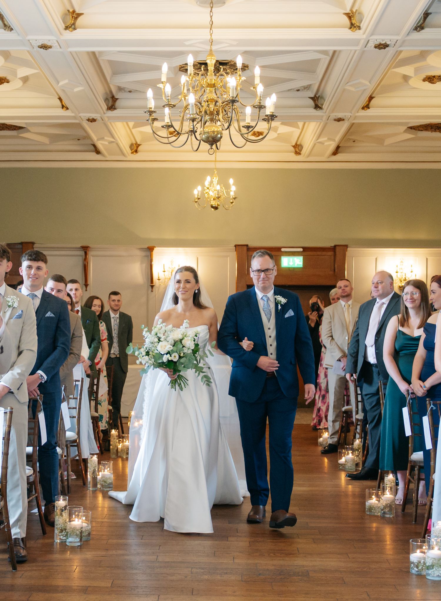 Elegant wedding ceremony in a grand ballroom with chandelier and candle-lined aisle.