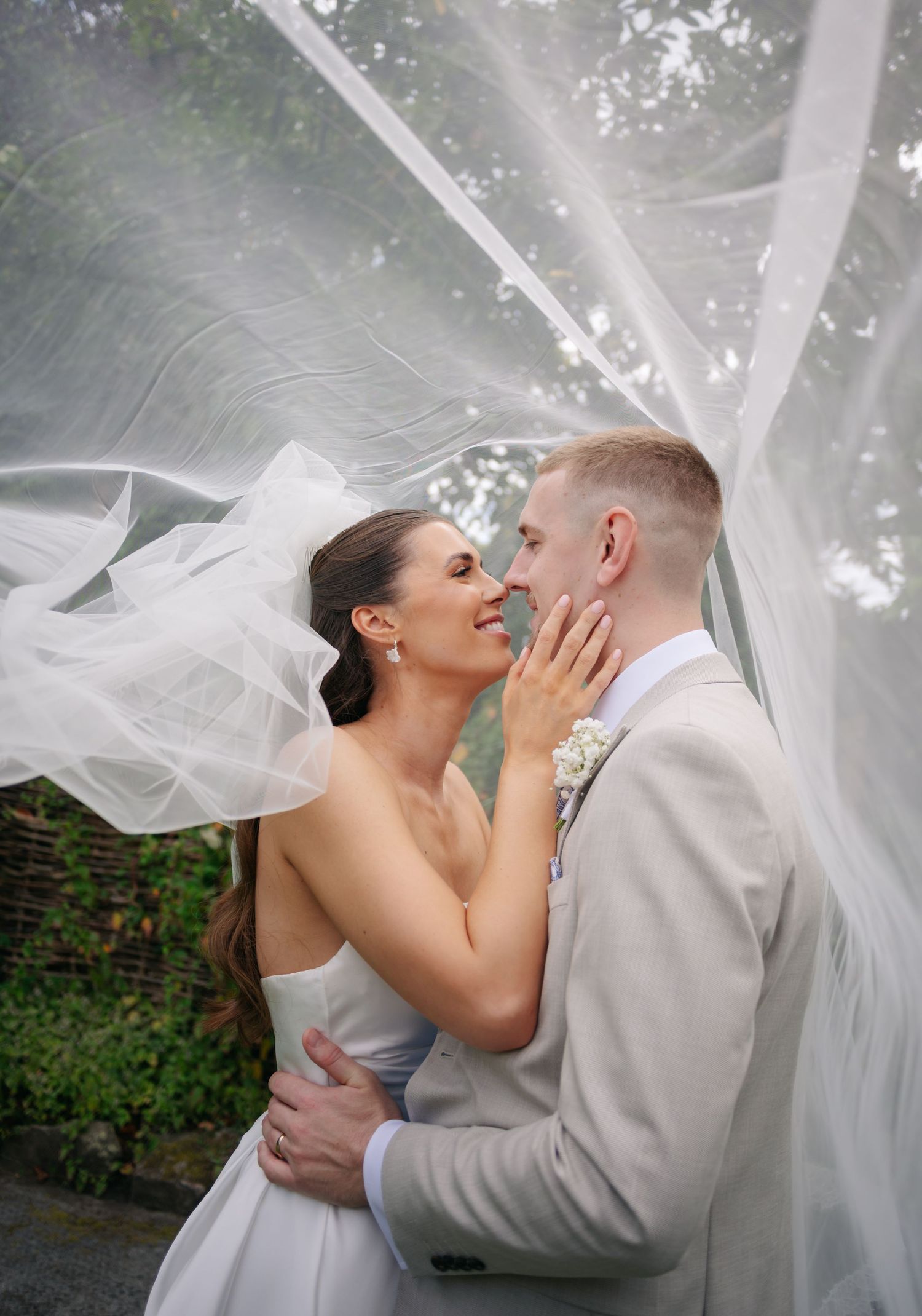 Romantic wedding couple embracing under flowing veil with dramatic lighting.