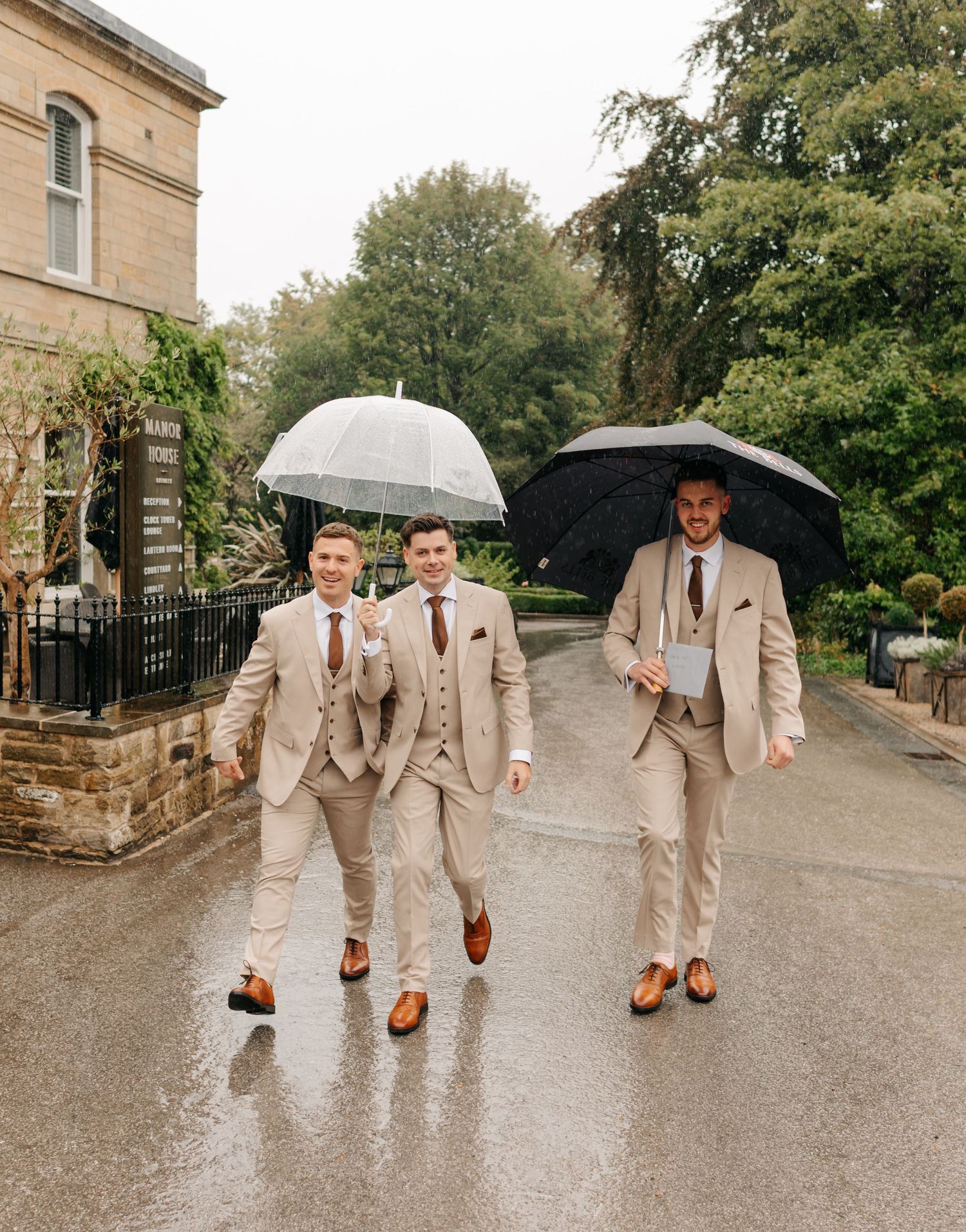 Groomsmen walking with umbrellas in tan suits on rainy wedding day.