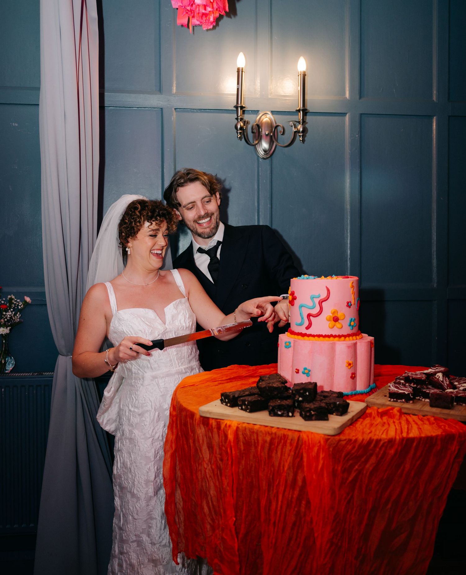 A couple cuts their pink wedding cake together at an elegant reception with candelabras and blue walls.