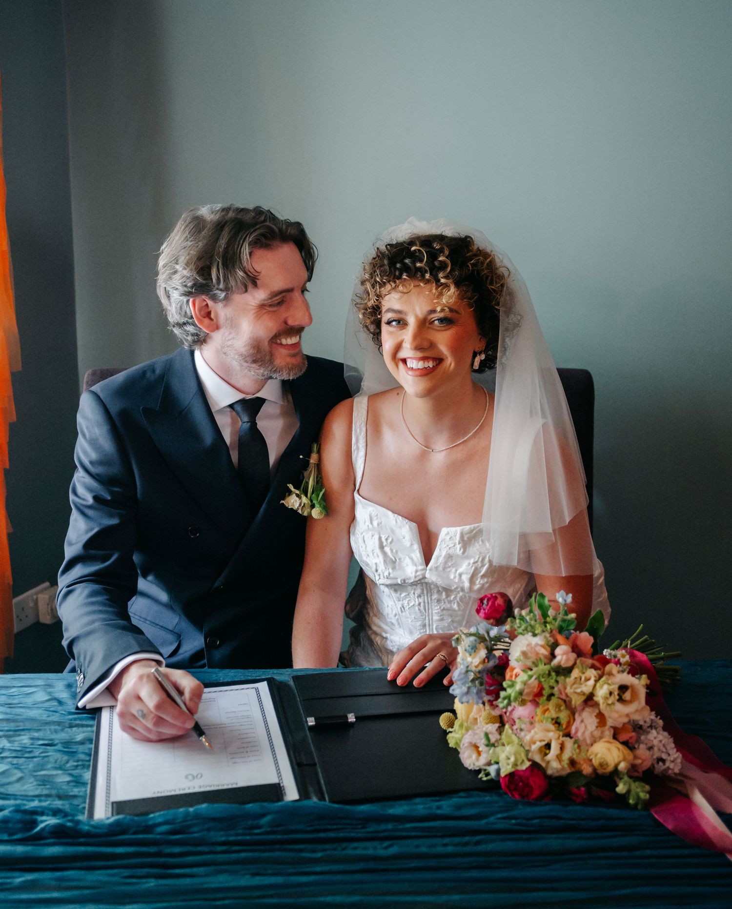 Couple signs marriage documents while sharing joyful moment at desk.