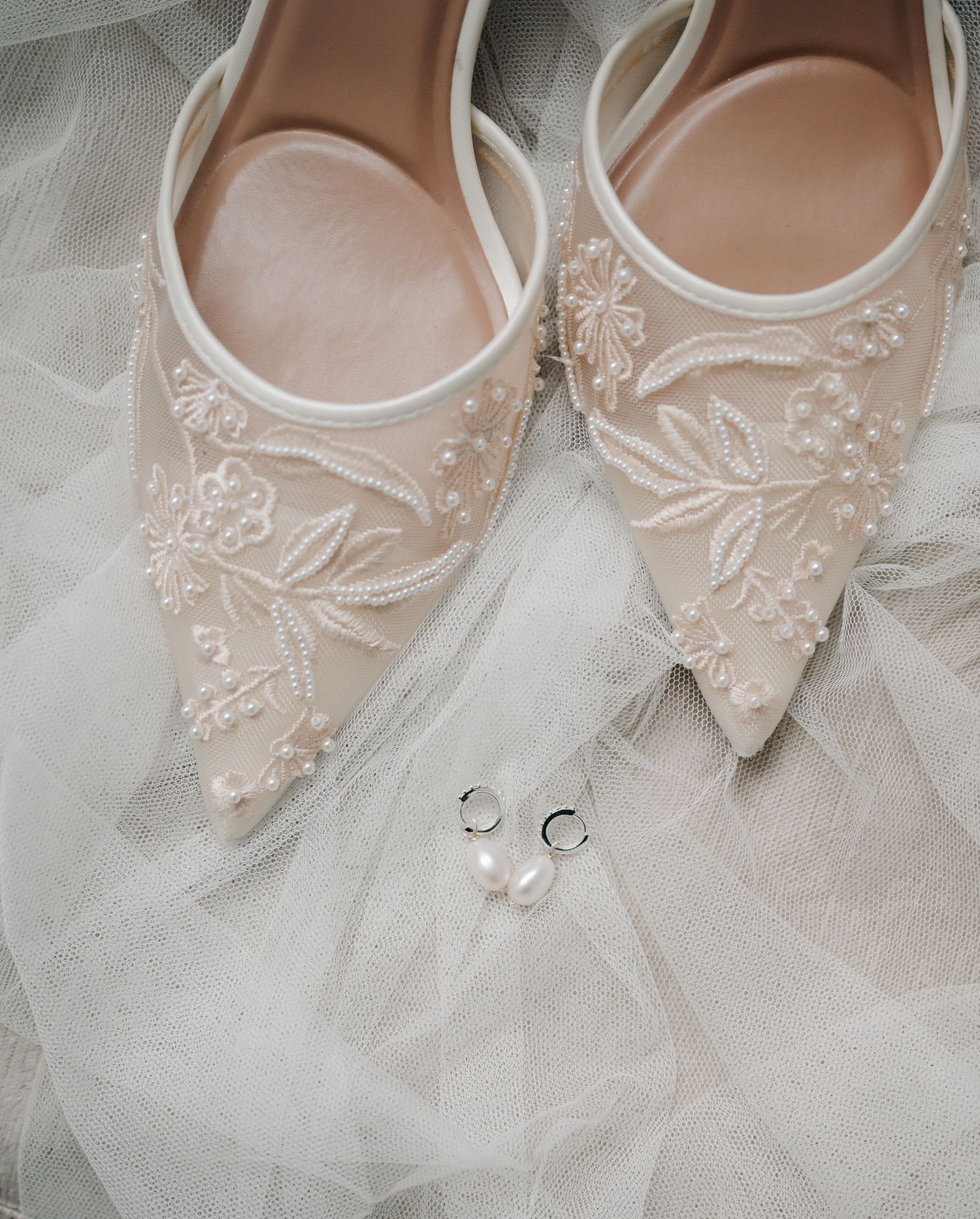 Elegant bridal shoes with delicate lace embroidery and pearl details displayed on white tulle fabric.