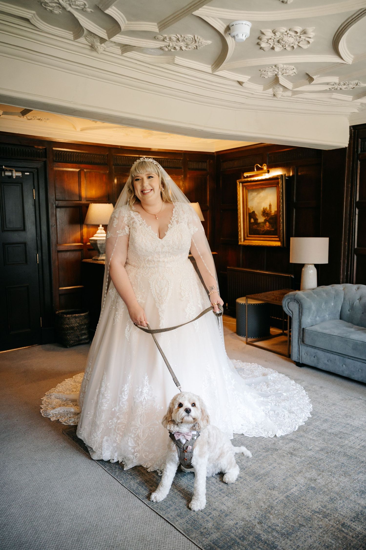 Smiling bride in lace gown holds leash of small dog in elegant wood-paneled room.