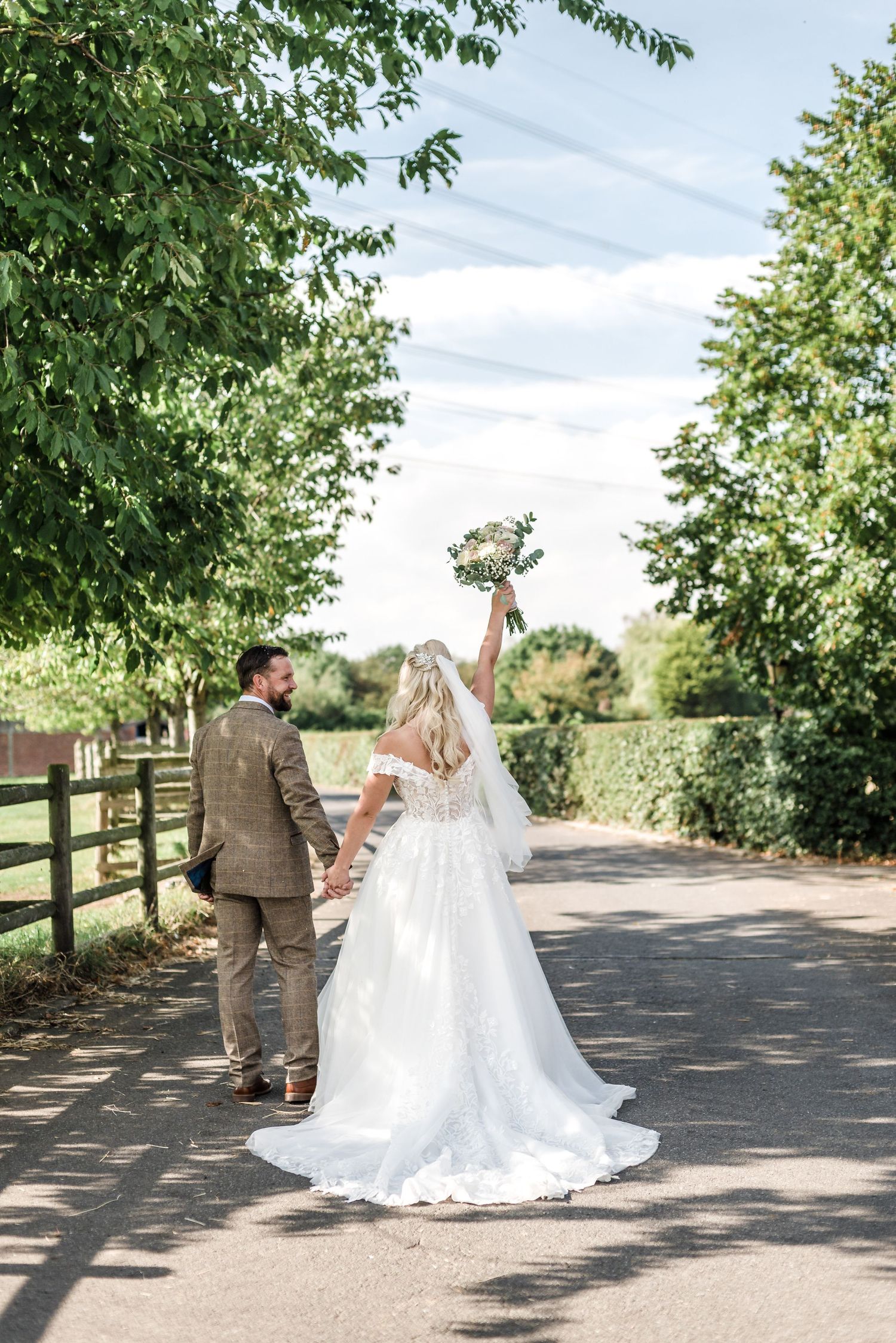 Bride in flowing white gown holds bouquet high while walking with groom on tree-lined path.