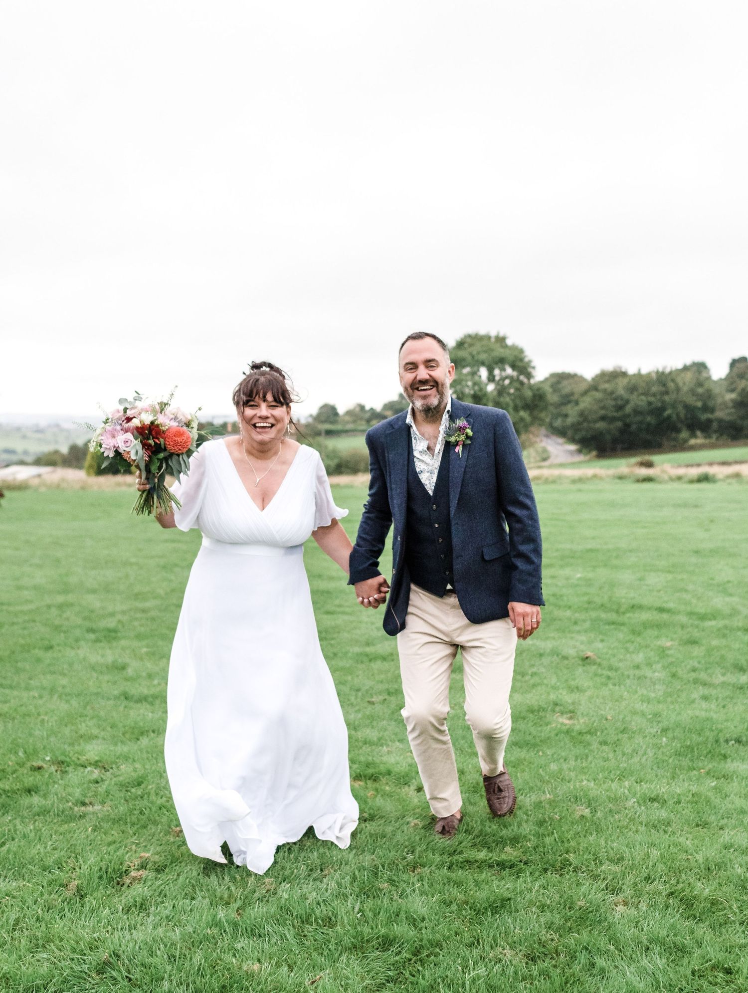 Joyful newlyweds walking hand in hand across a green field on their wedding day.
