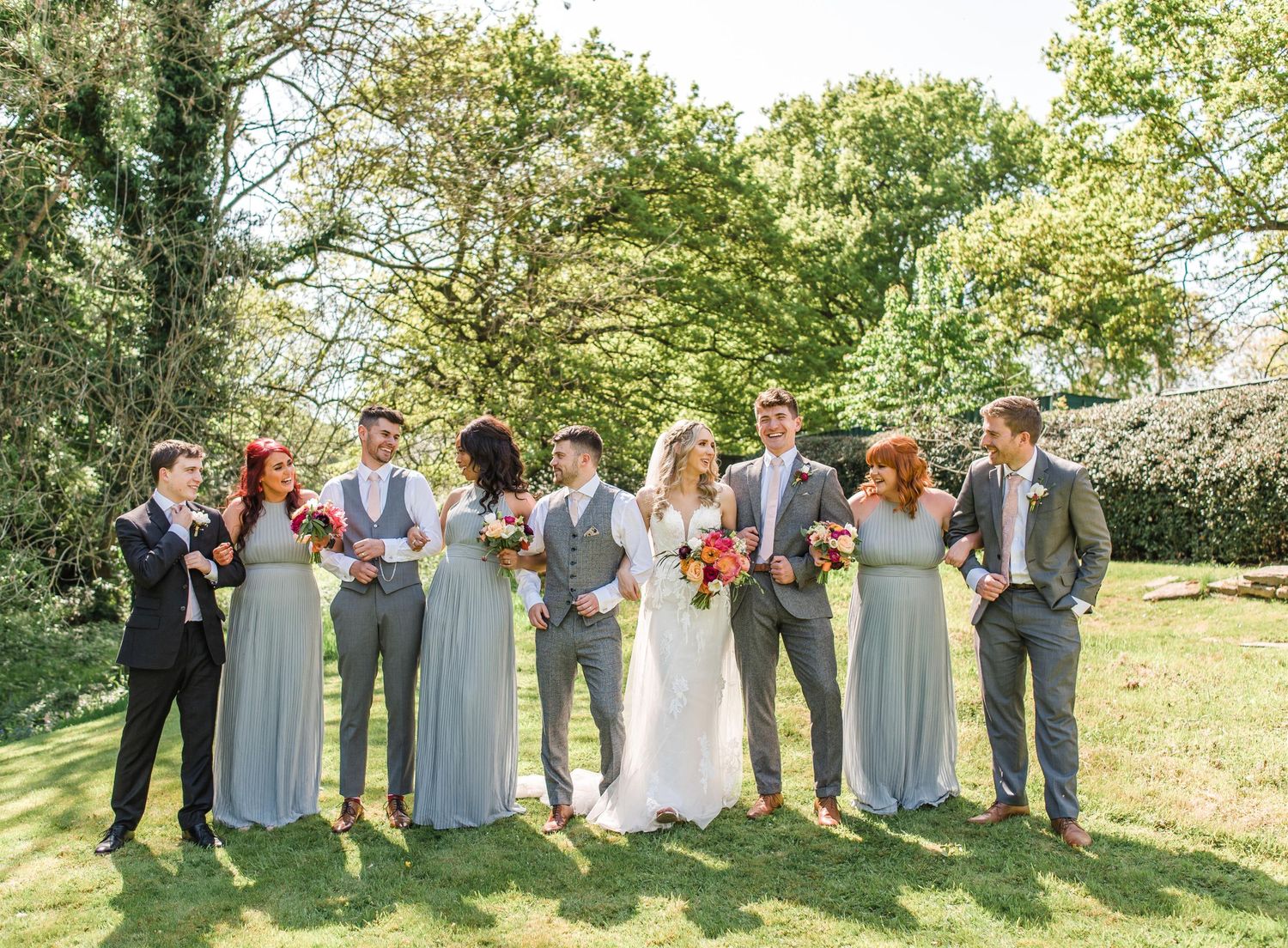 Full wedding party in sage green and gray attire poses together outdoors.