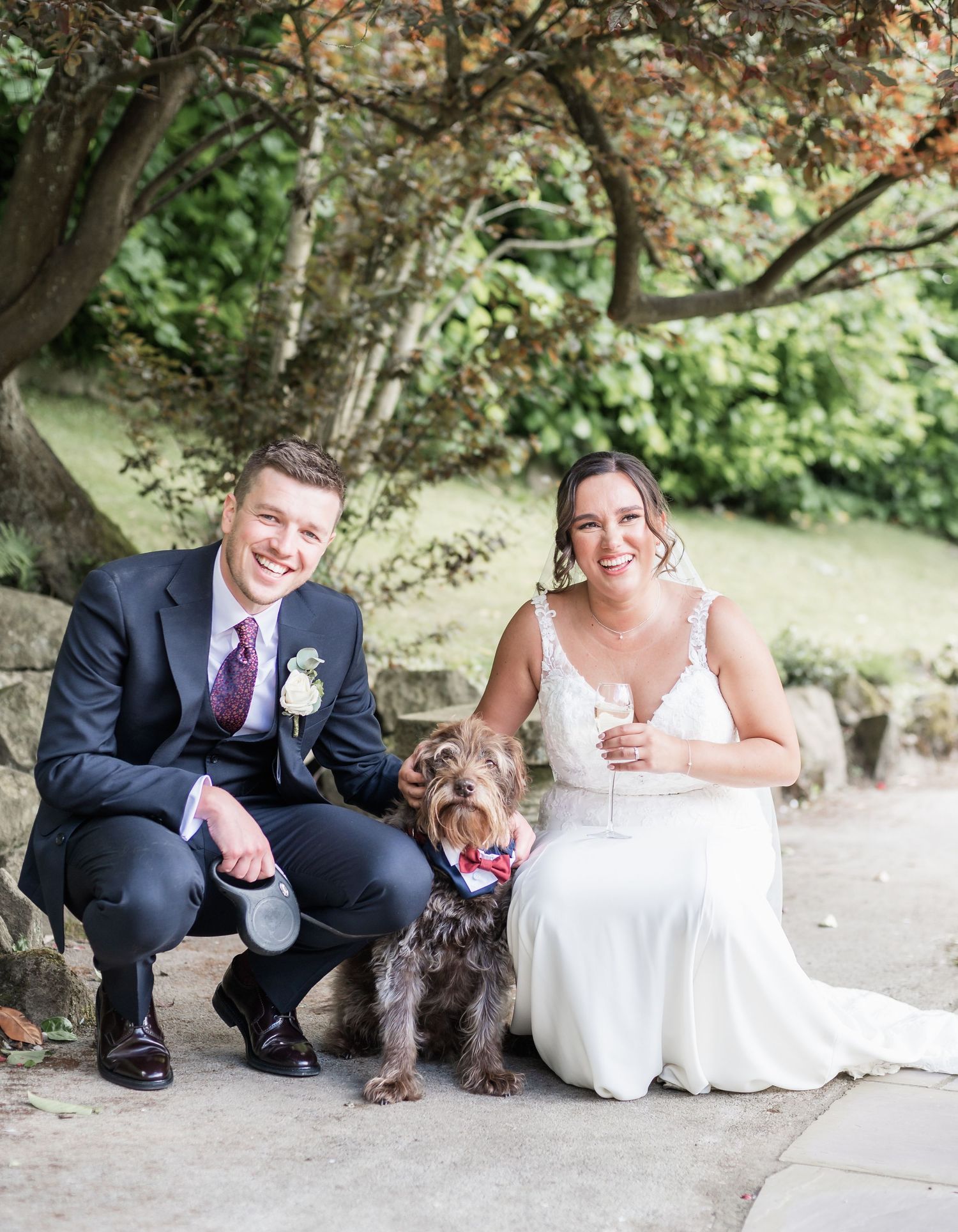Wedding couple poses with their small brown dog on garden pathway.