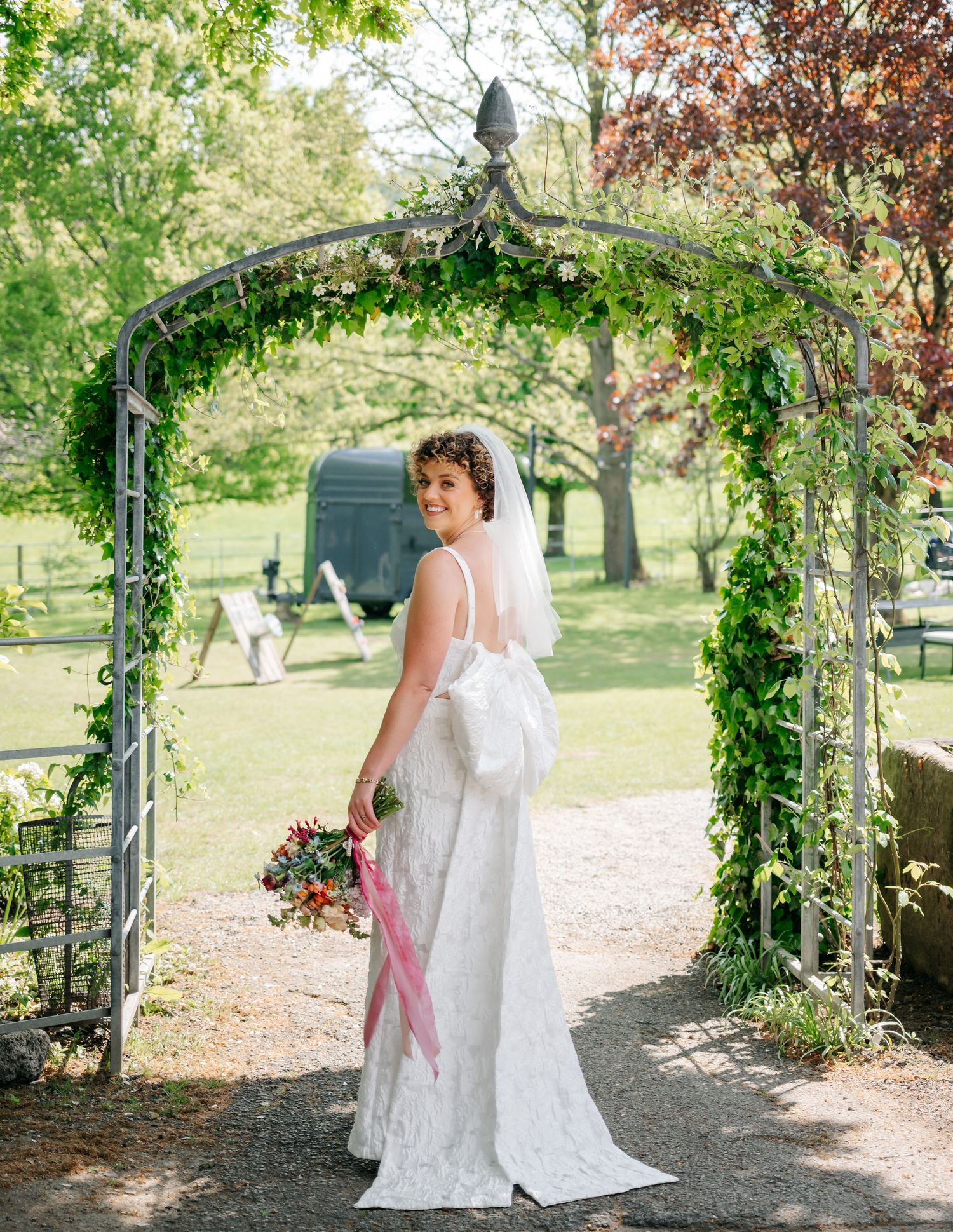 Lace-dressed figure poses beneath a greenery-covered garden arch on a sunny day.