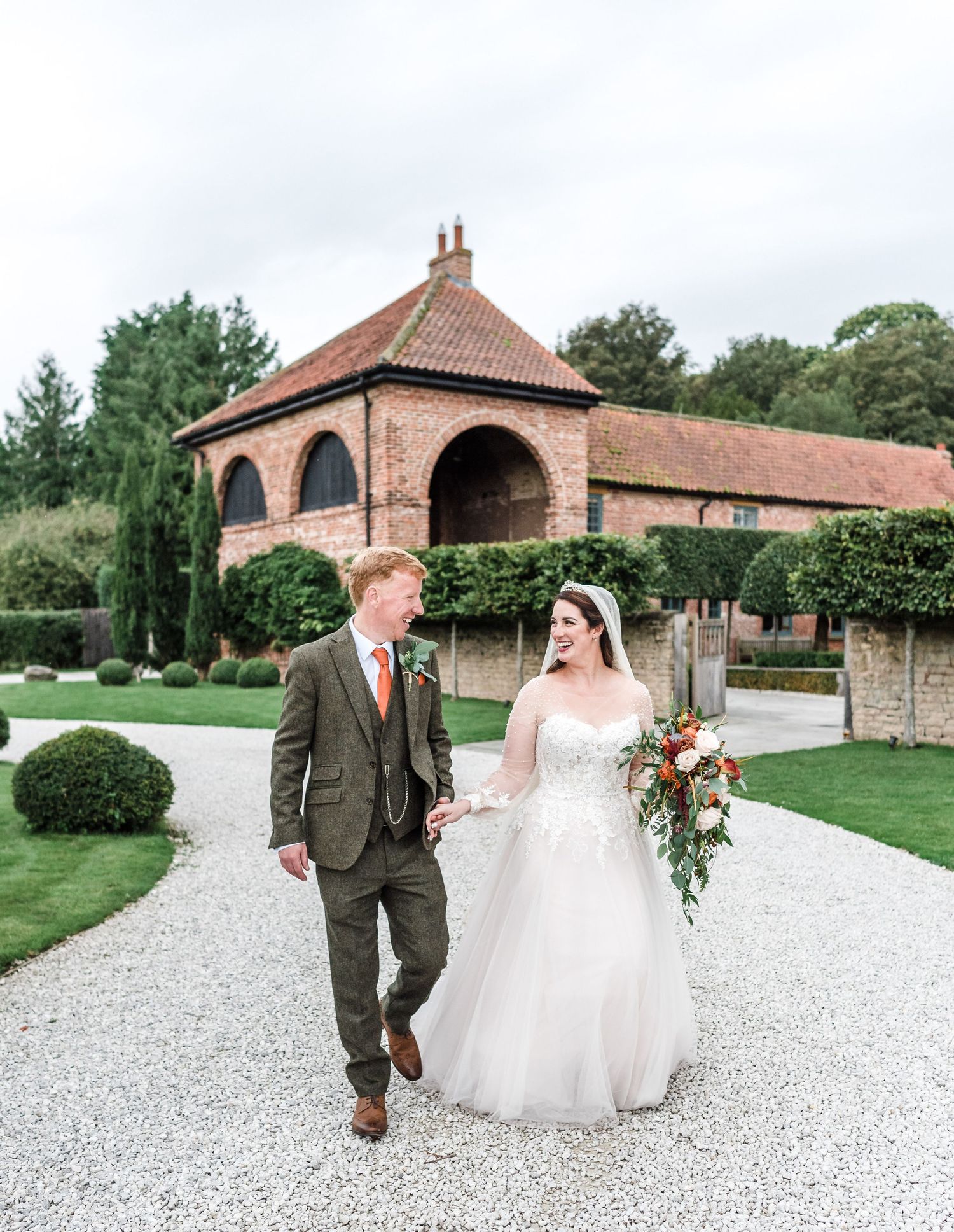 Newlyweds walk together on gravel path in front of brick archway building with manicured garden.