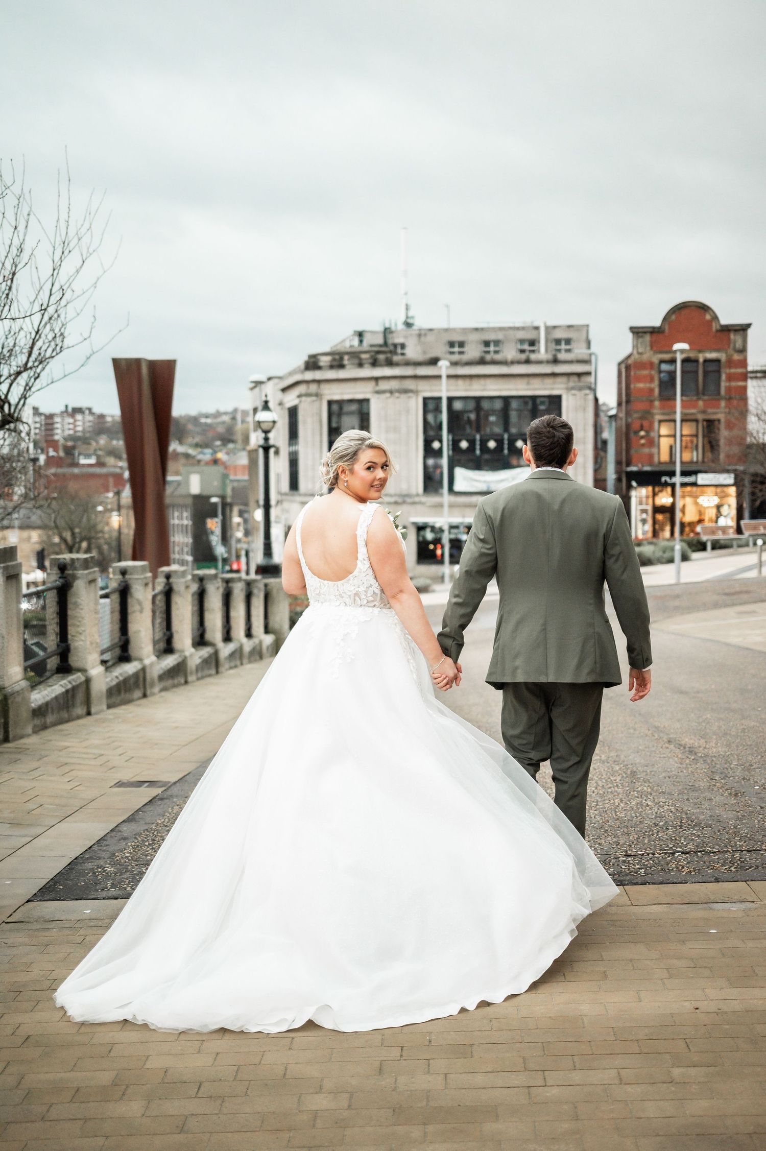 A wedding couple walks along a city street with historic buildings and a rustic metal sculpture in the background.