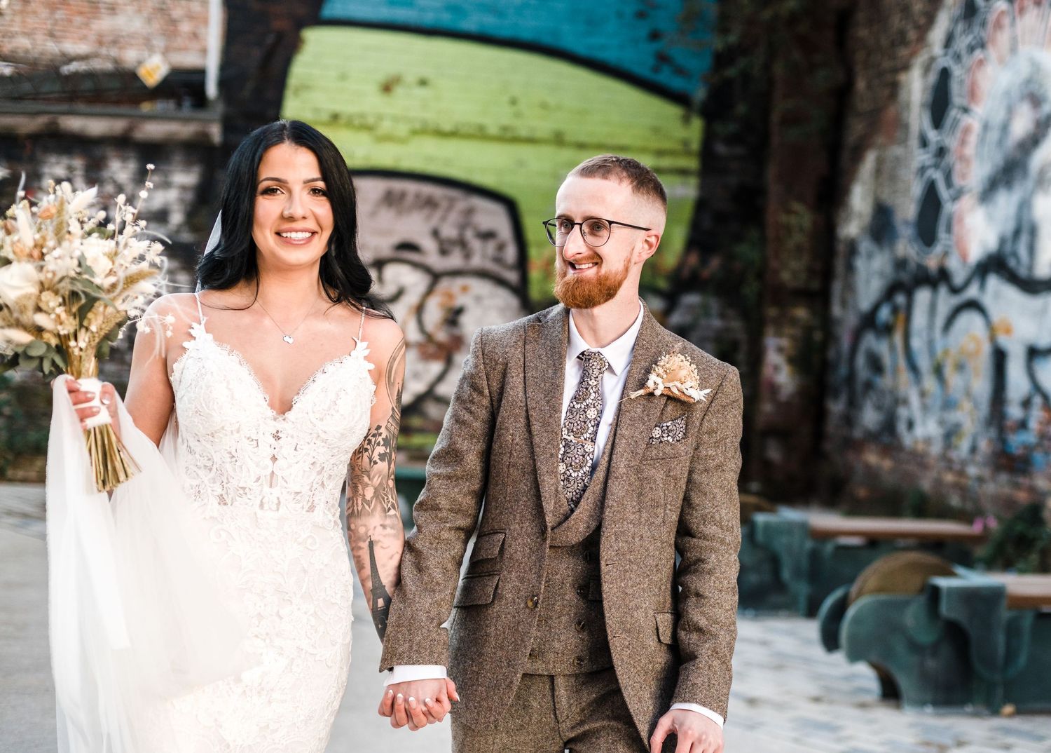 Newlyweds hold hands and smile while standing in urban graffiti setting.