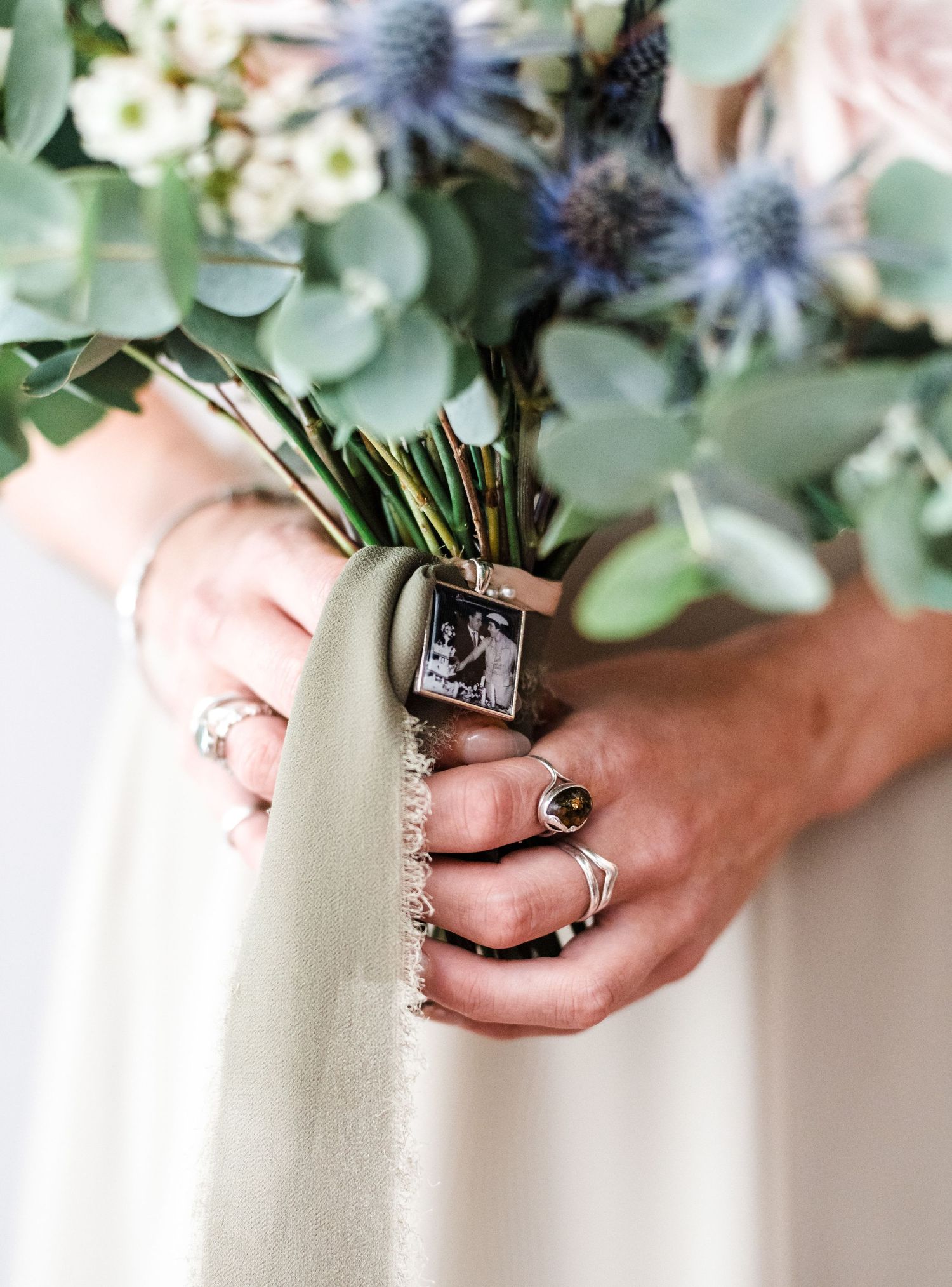 Close-up of hands holding wedding bouquet with sentimental photo charm and vintage rings.