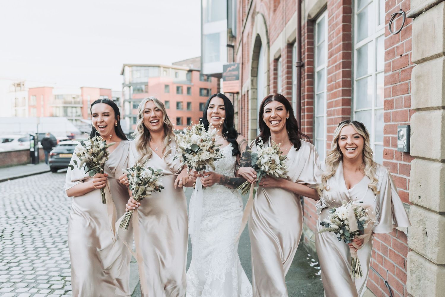 A bridal party wearing neutral toned dresses poses for photos along a brick wall and cobblestone walkway.