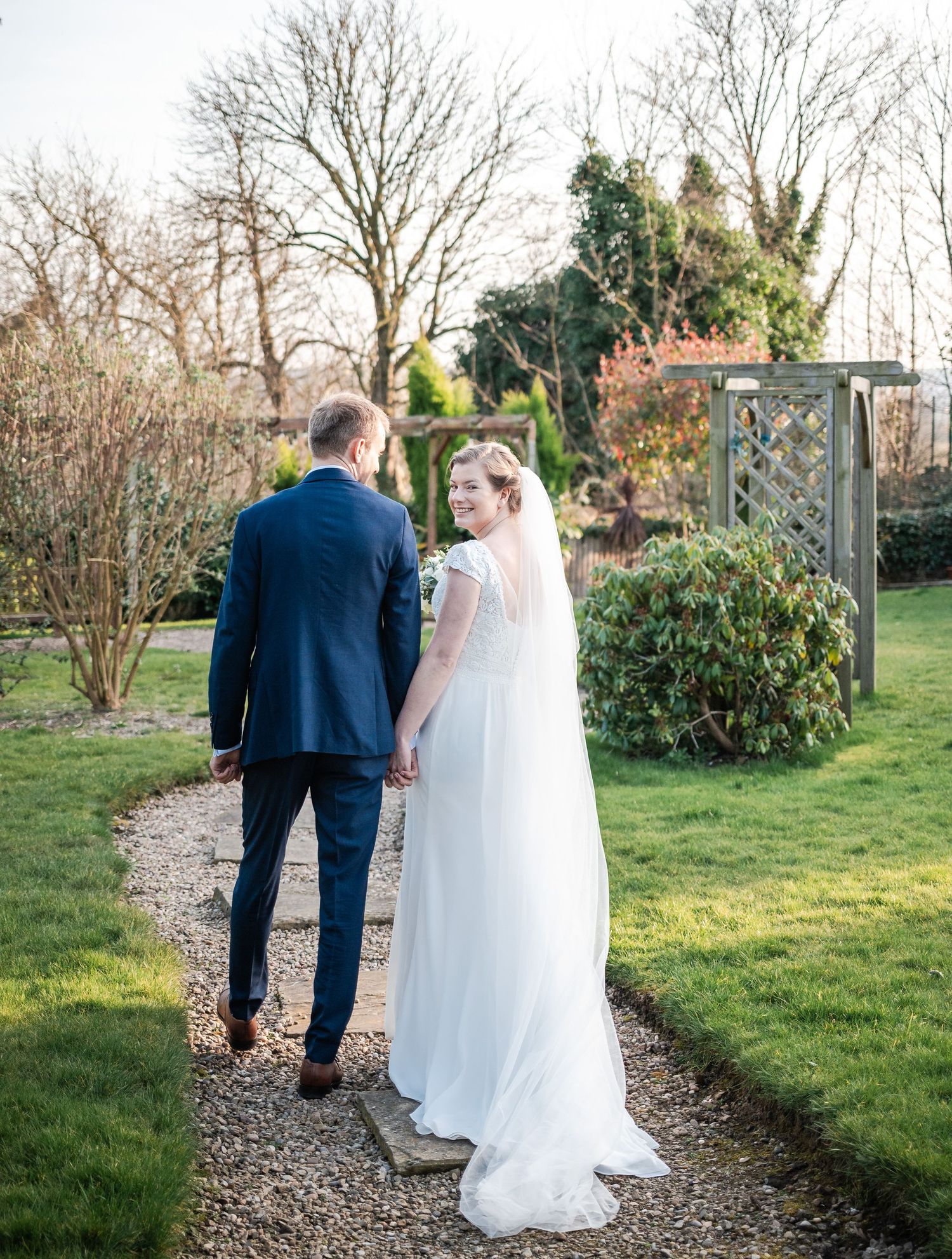 Newlyweds walk along a garden path surrounded by winter trees and green shrubs.