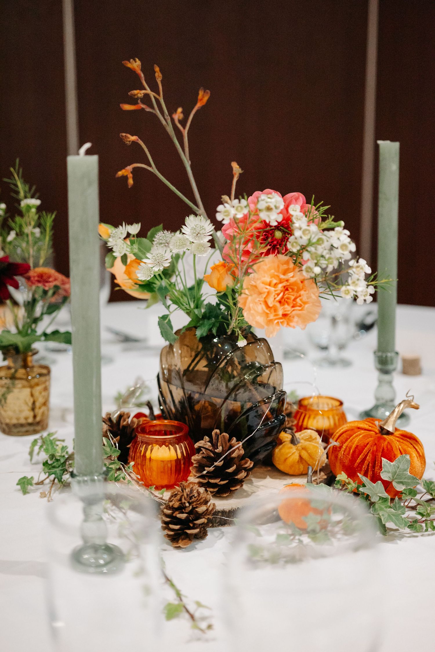 Fall-themed table centerpiece with orange flowers, pine cones, and pumpkin decorations.