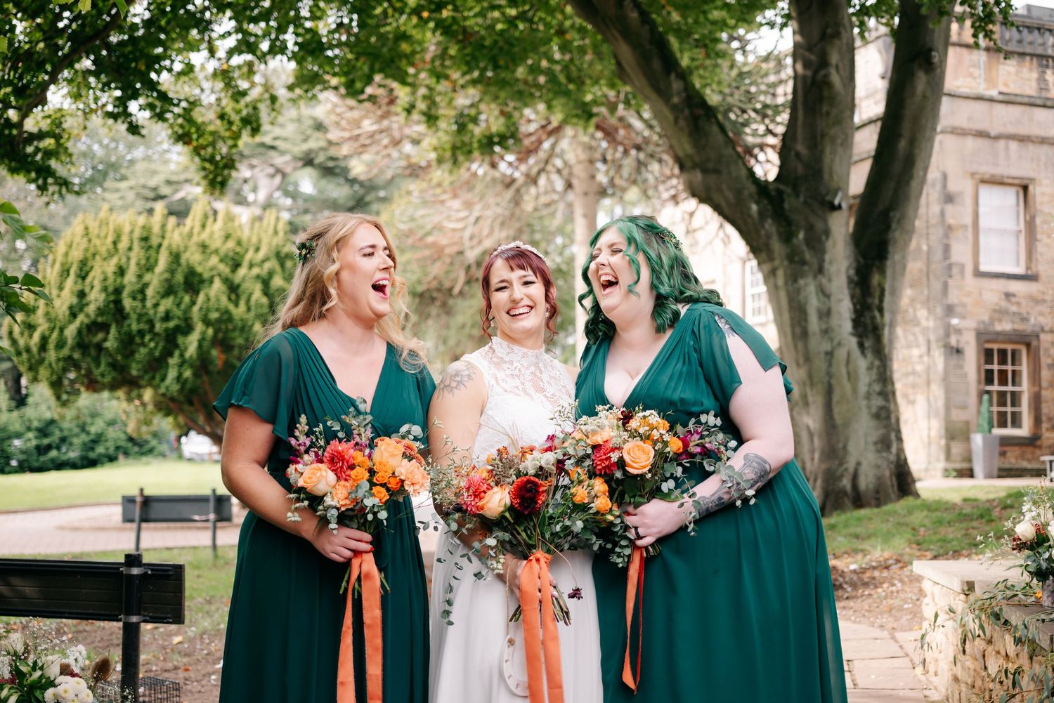 Bridesmaids in emerald green dresses posing outdoors under mature trees.