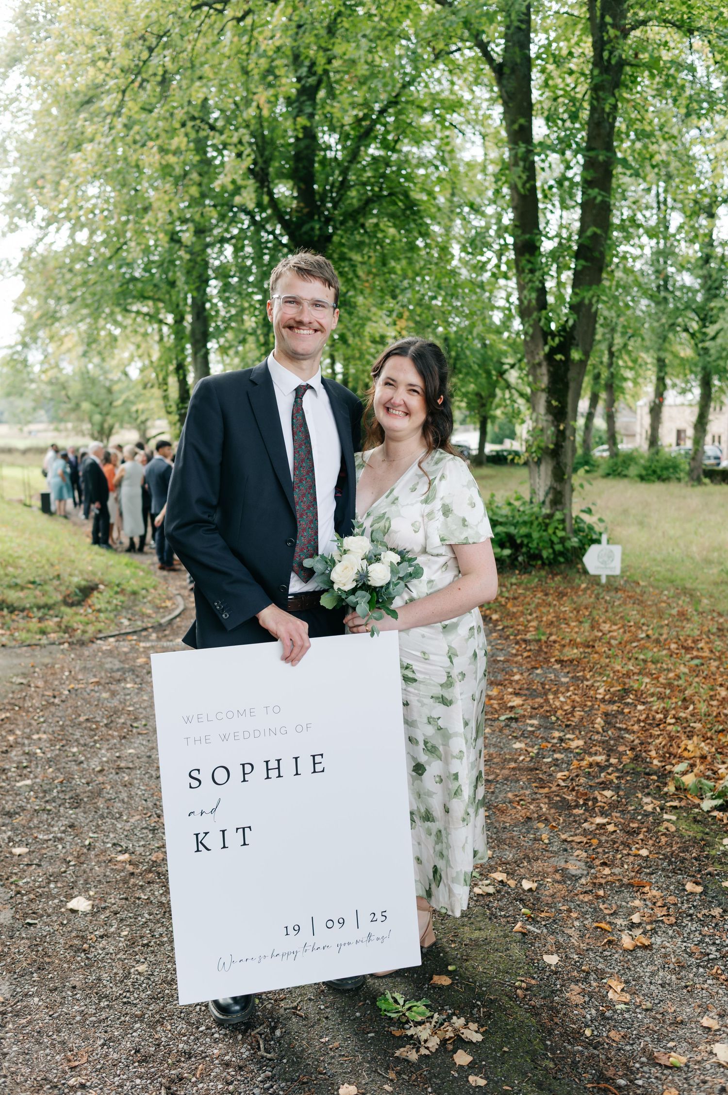 A couple poses outdoors in autumn holding signs on their wedding day among fallen leaves and trees.