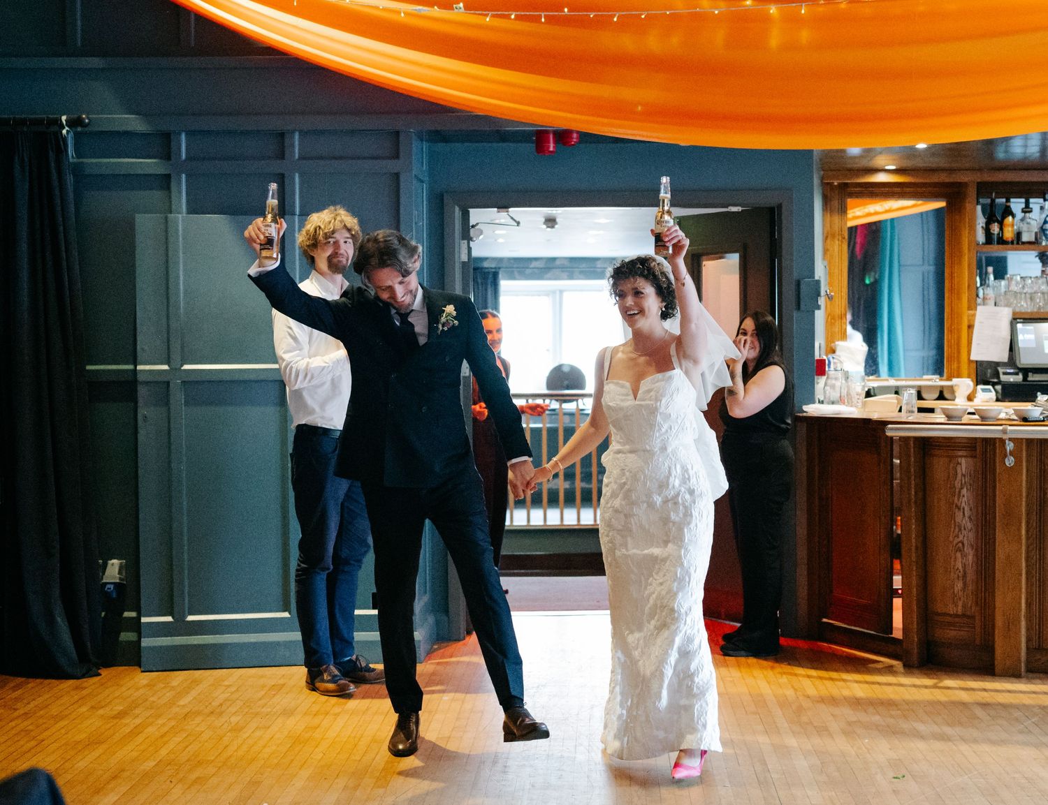 A wedding reception venue with orange draping overhead and dark blue walls features couples dancing on a wooden floor.