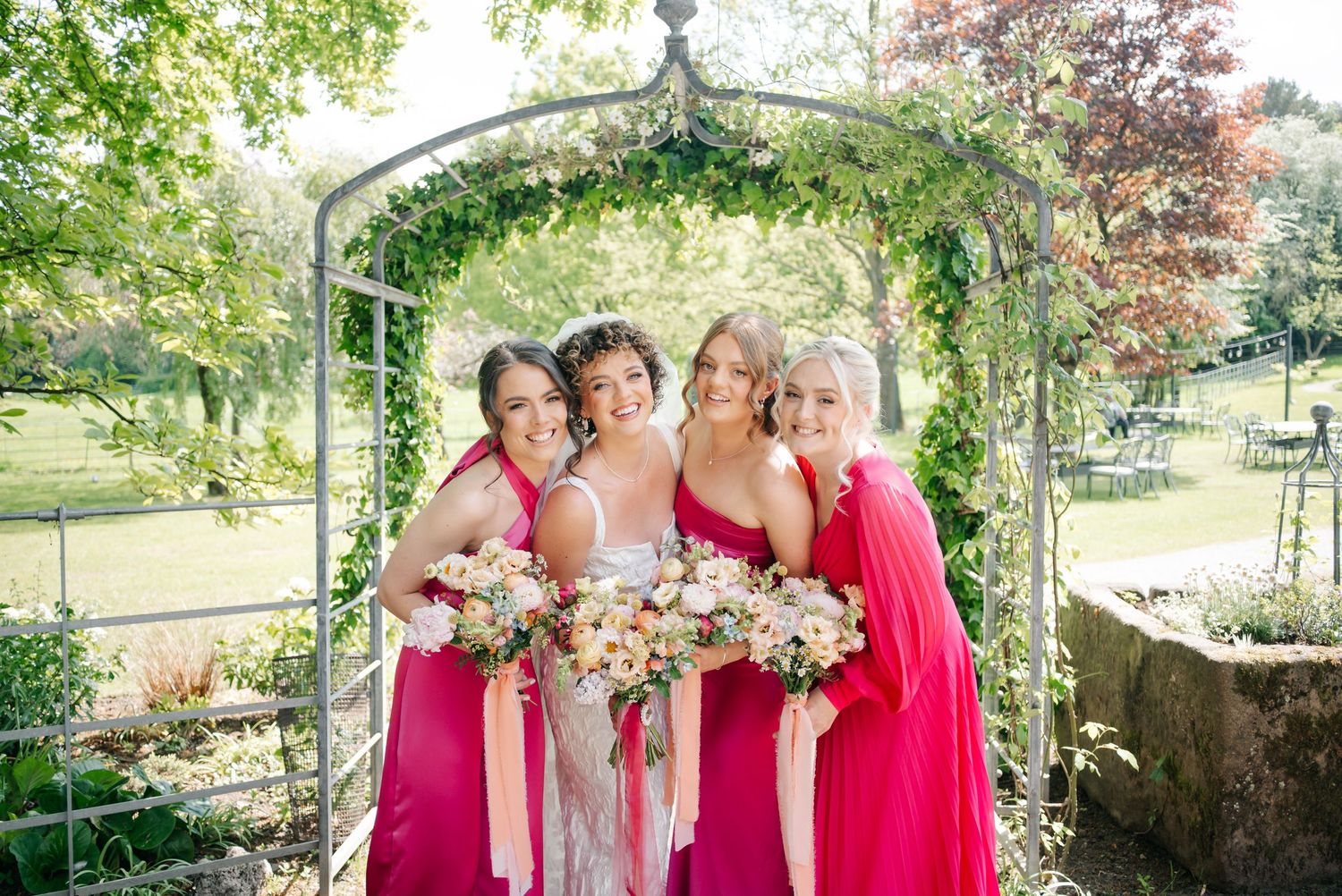 Bridesmaids in bright pink dresses pose under a floral garden arch.