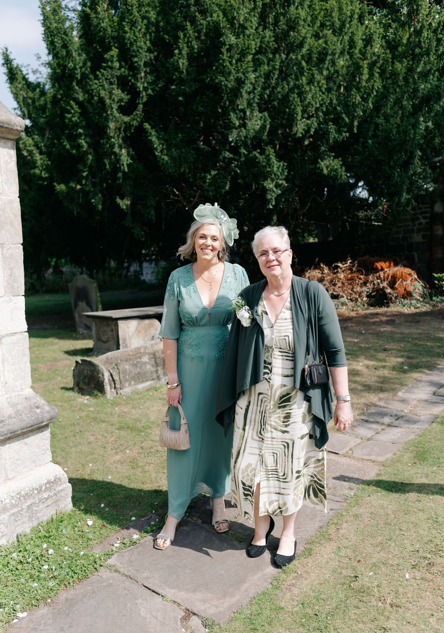 Wedding guests in green and patterned dresses pose together on church grounds.