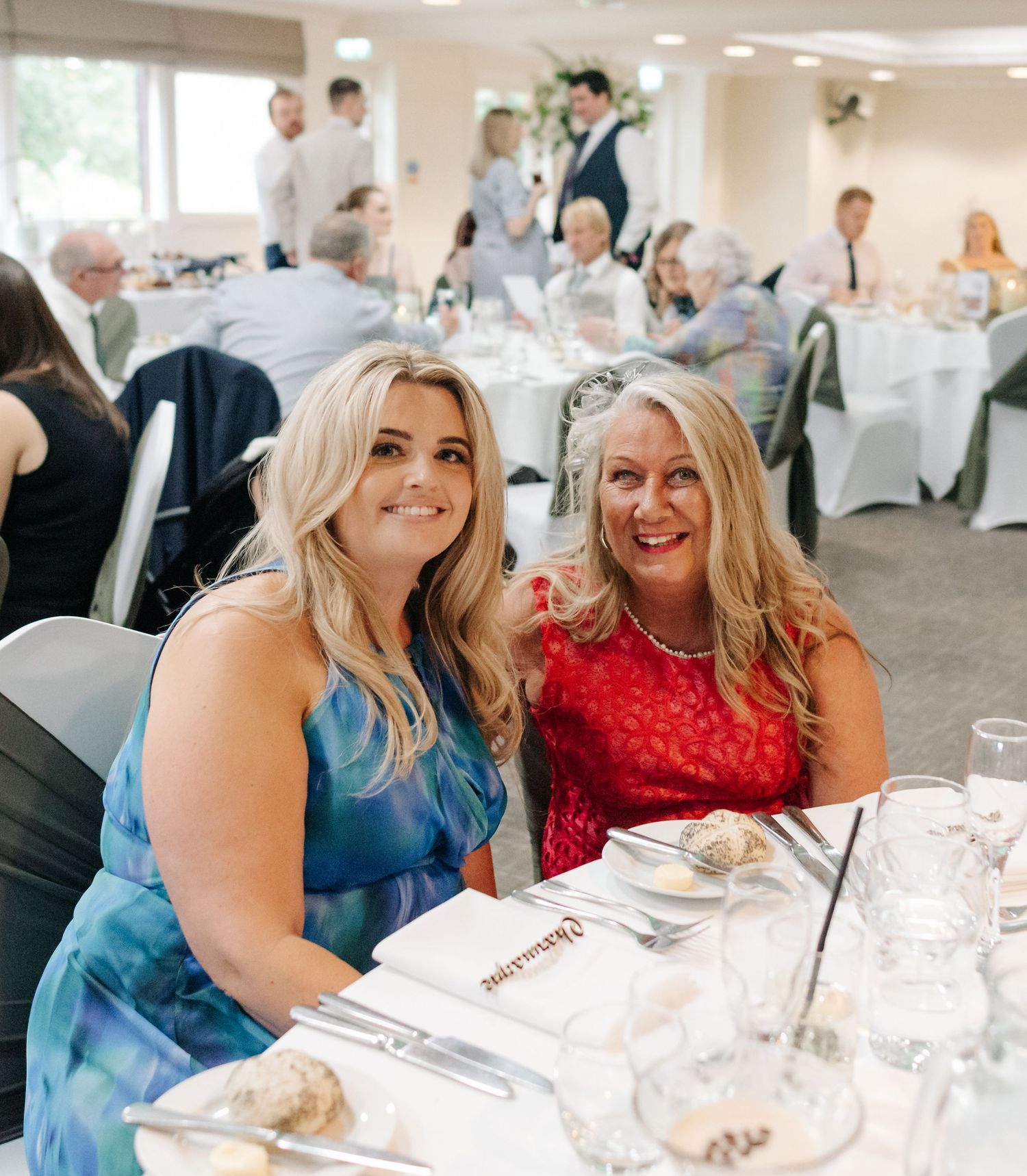 Wedding guests in blue and red dresses laugh together at reception tables.