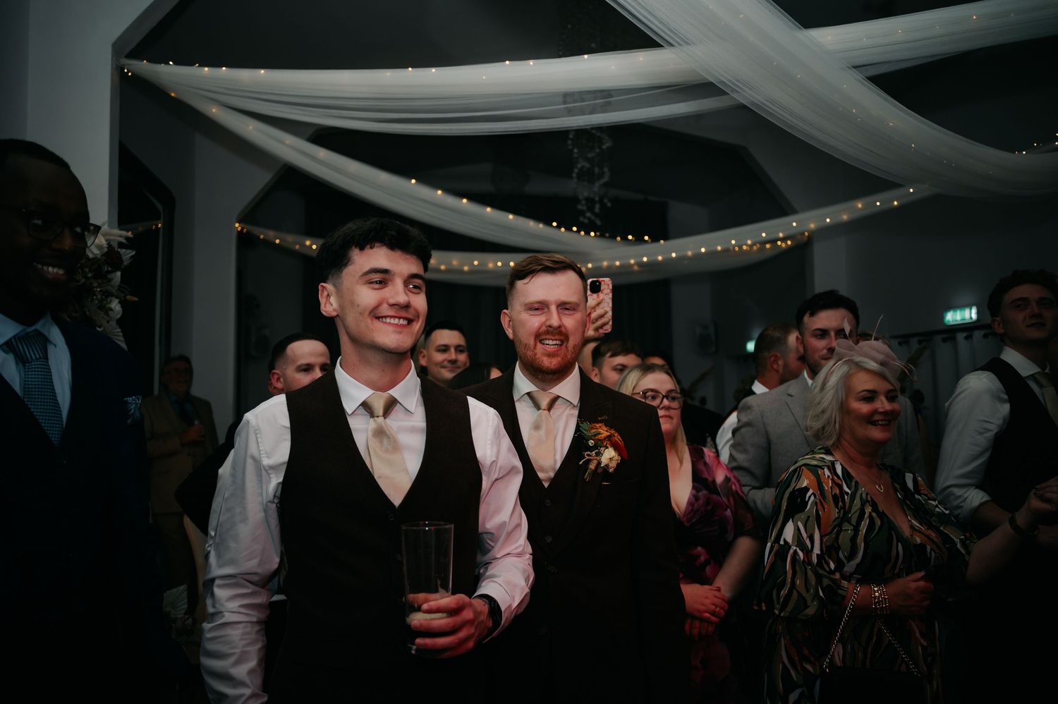 Wedding reception party scene with guests celebrating under white draped ceiling with string lights.