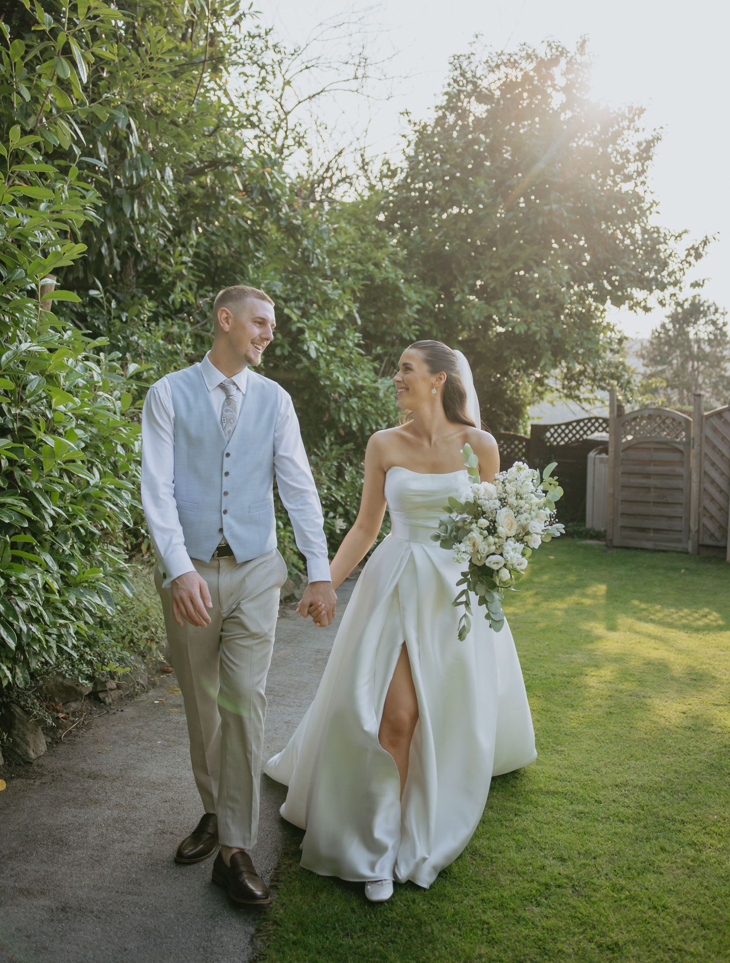 Couple walks through a sunlit garden path on their wedding day with flowing white dress.