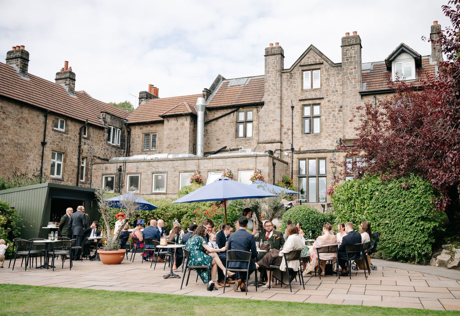 Wedding guests sit at tables and under blue parasols on a sunny stone terrace outside a historic manor venue.
