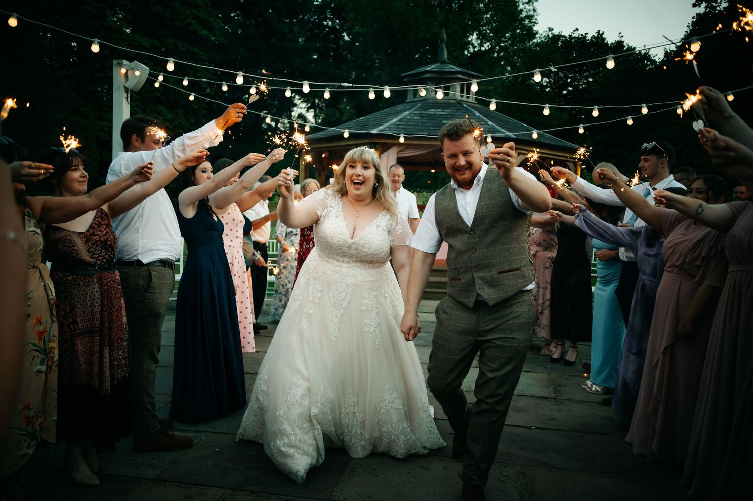 Newlyweds walk through sparkler tunnel with cheering guests under fairy lights at magical evening wedding reception.