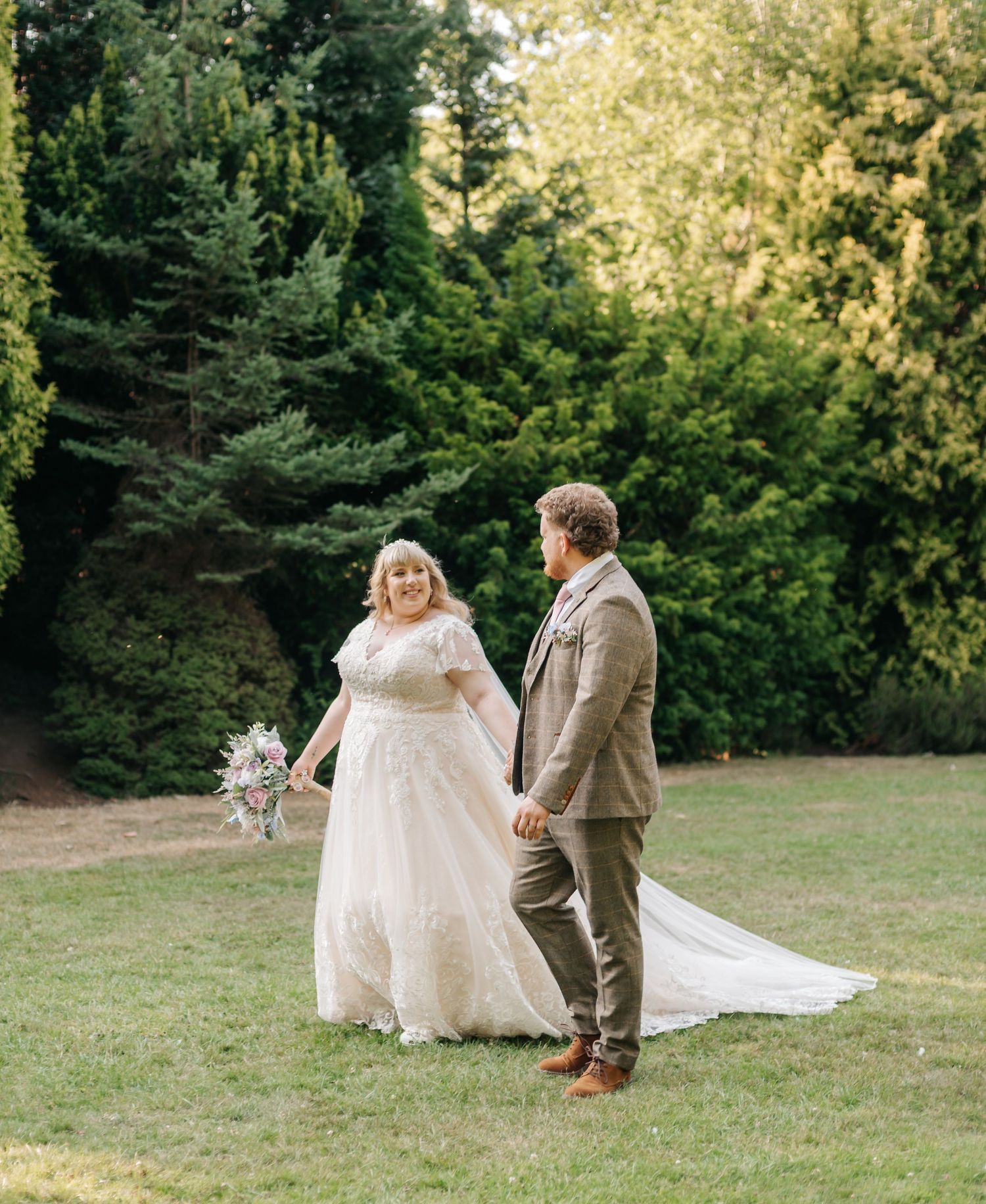 A bride in a flowing white gown holds a bouquet while walking hand-in-hand with a groom in a brown suit on a green lawn.