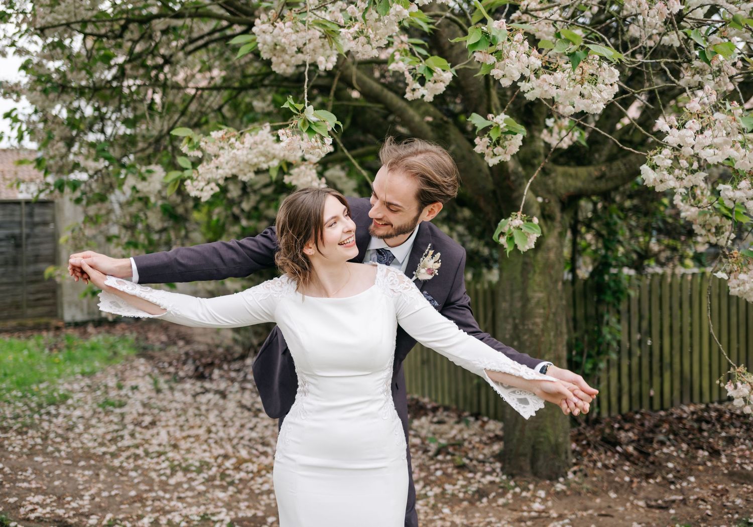 Wedding couple poses beneath blooming spring trees in an outdoor garden setting.