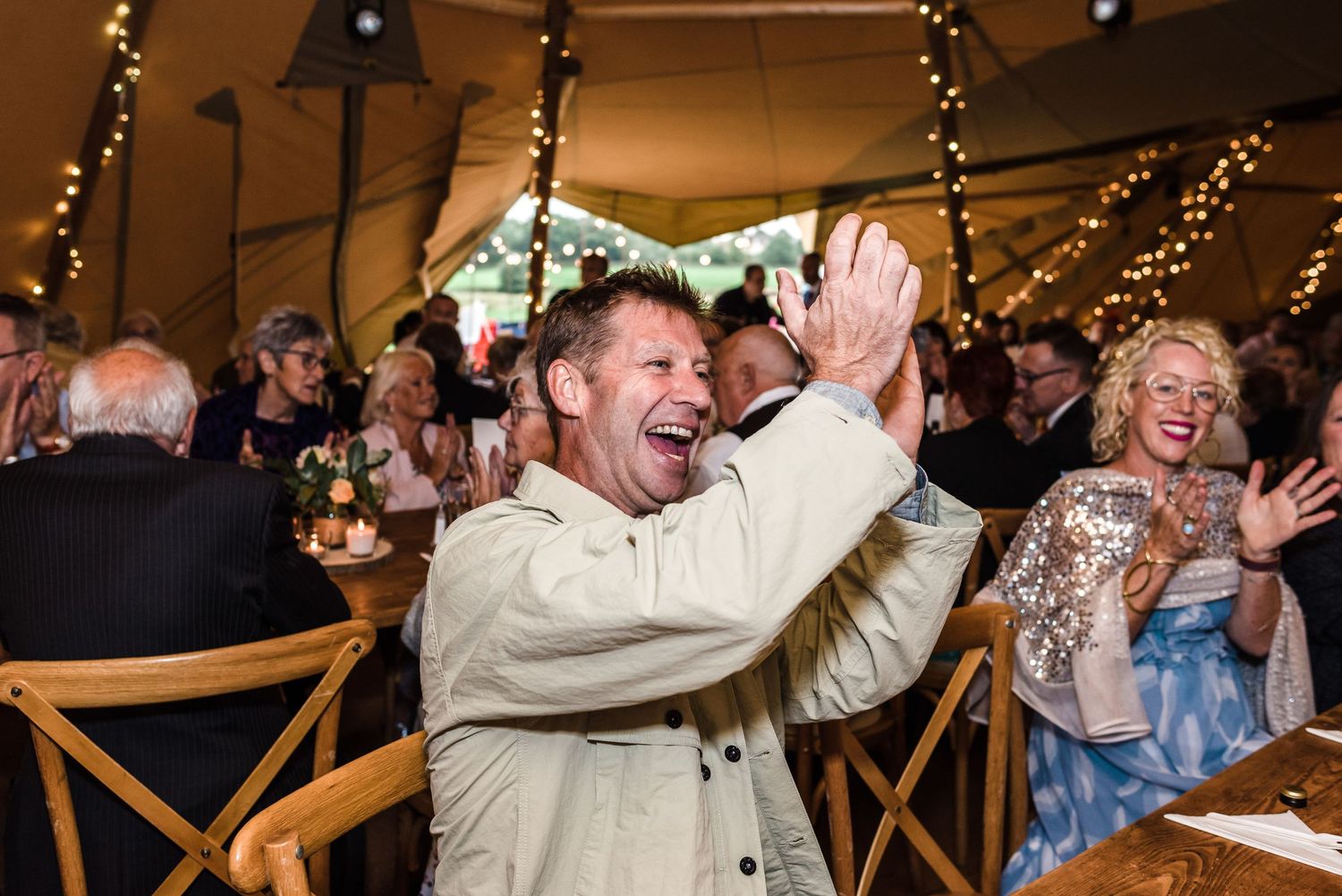 Wedding guests celebrating joyfully inside a tent with string lights and wooden chairs at the reception.