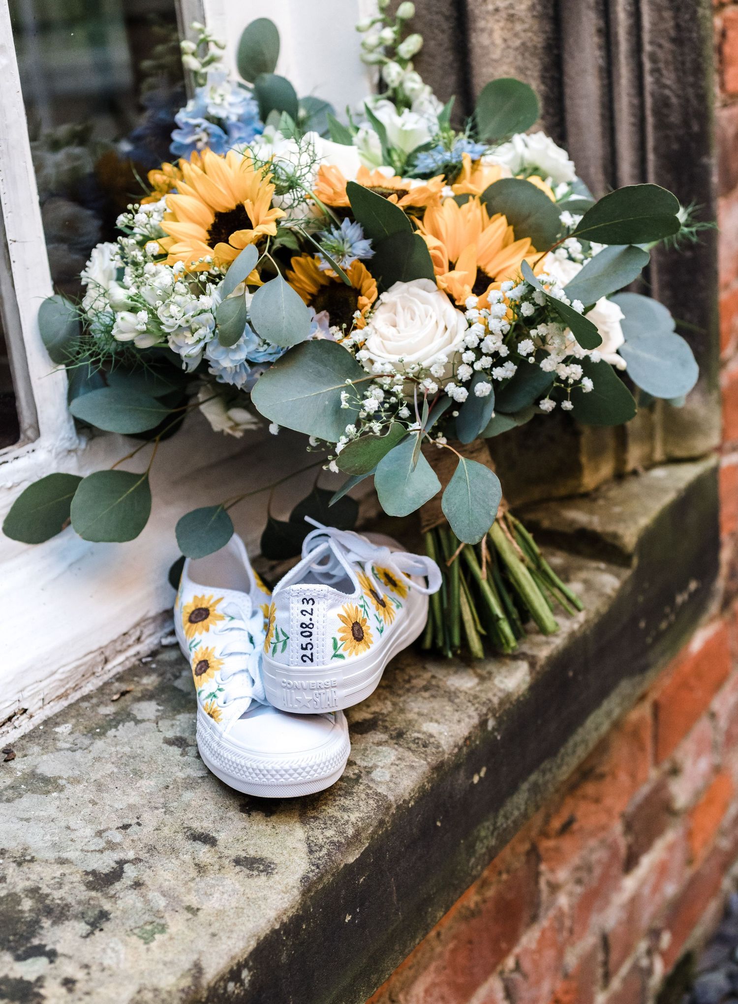 Bridal bouquet with sunflowers and white flowers next to decorated wedding sneakers on stone ledge.