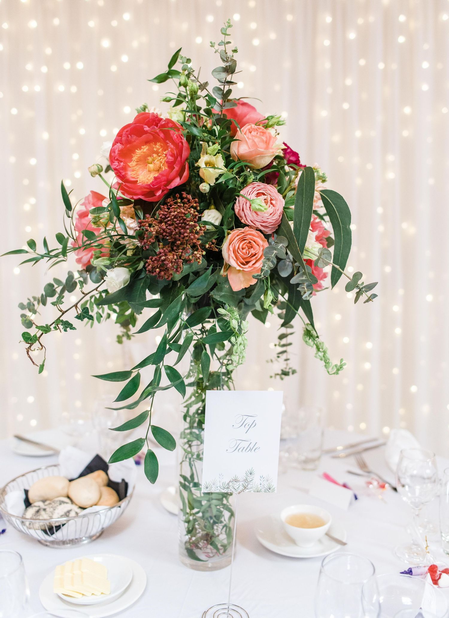 Elegant coral and pink floral centerpiece with greenery against twinkle light backdrop.