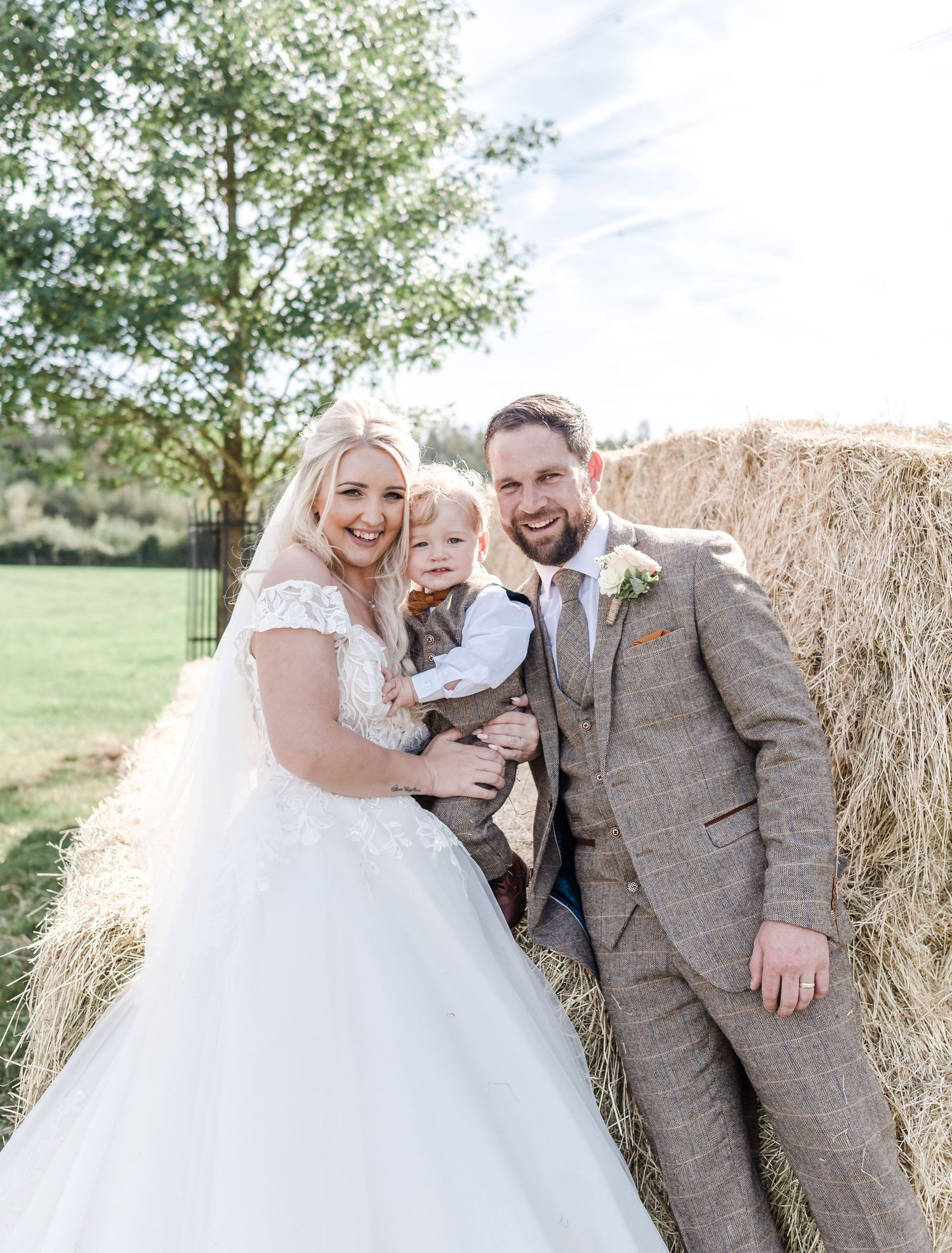 Wedding portrait of a couple and small child posed against a hay bale on a sunny day.