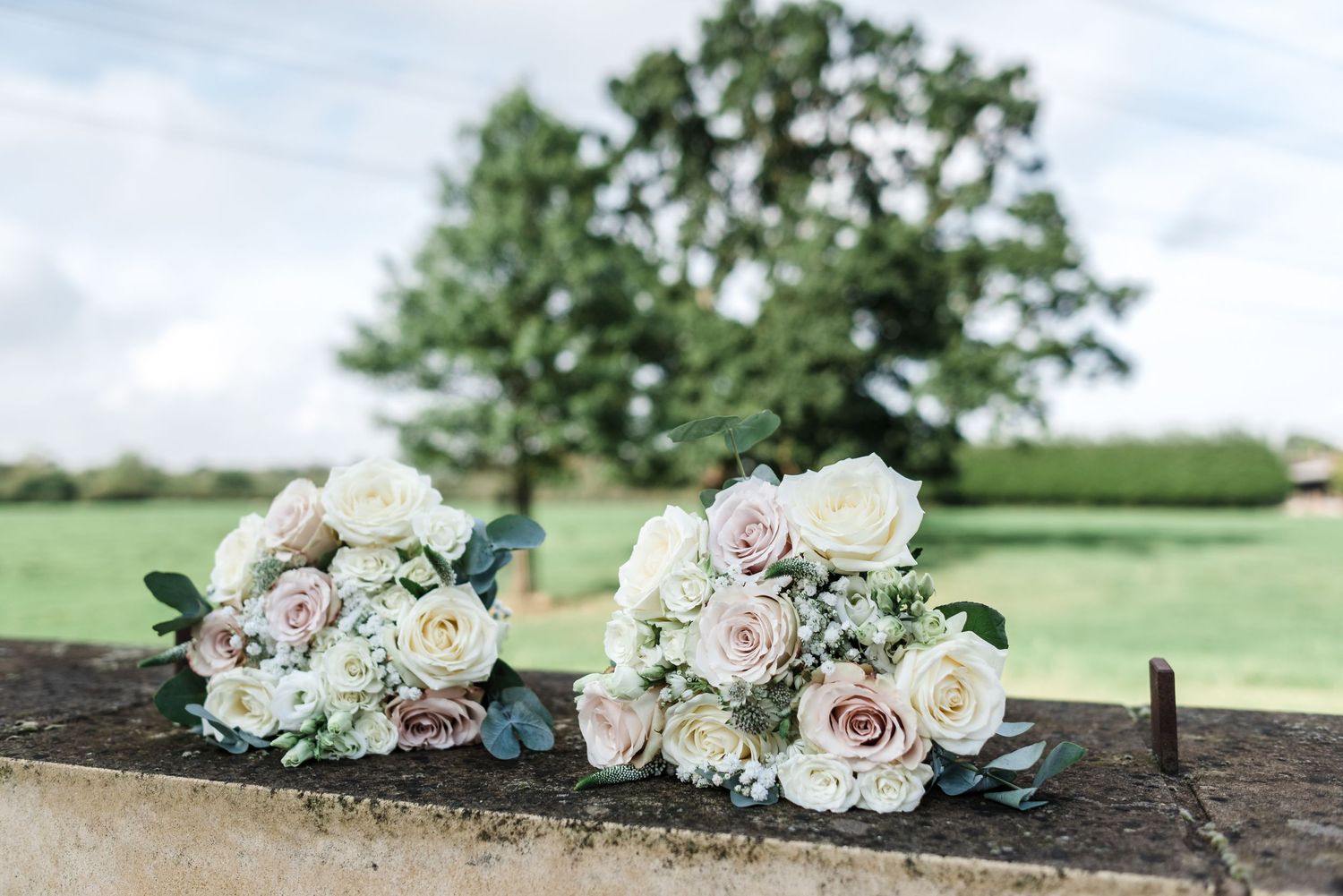Two delicate wedding bouquets with pink and white roses resting on stone wall with green countryside backdrop.
