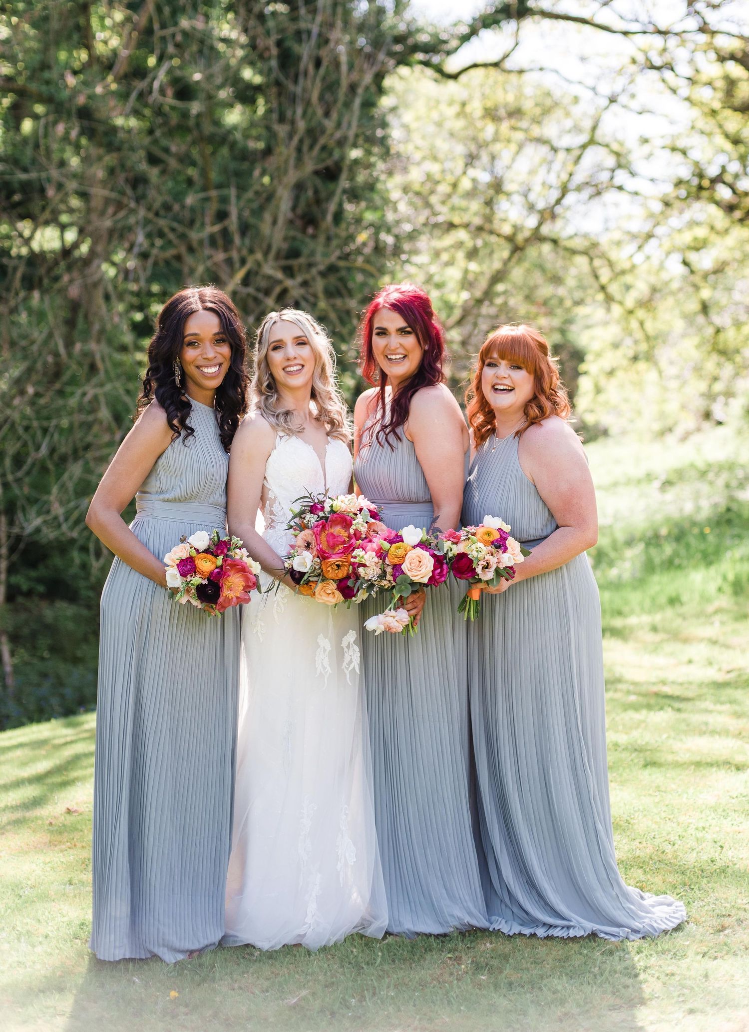 Bridal party in flowing gray dresses holding vibrant bouquets in garden setting.