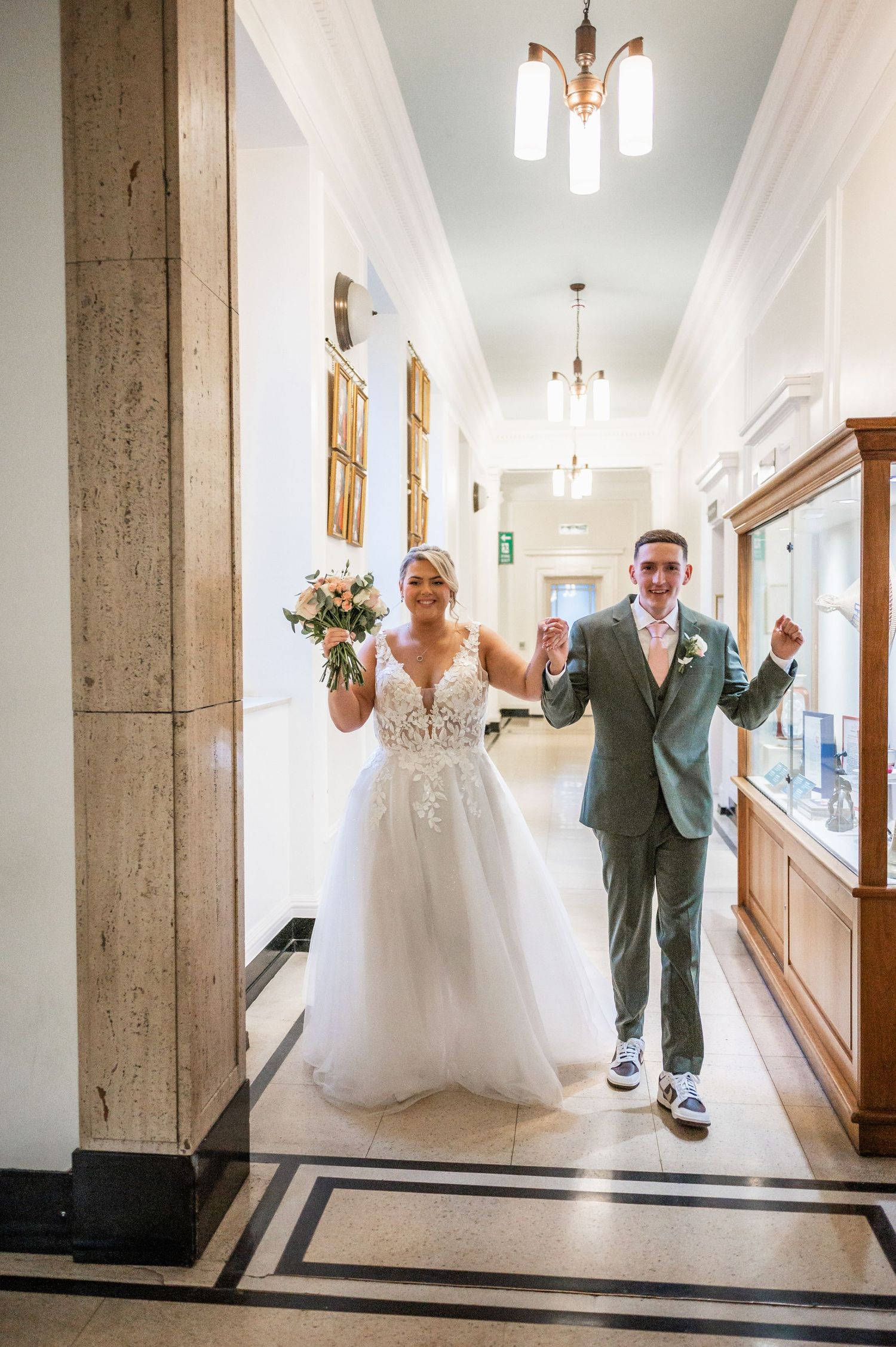 A happy couple celebrating their marriage walks down an elegant hallway with chandelier lighting.
