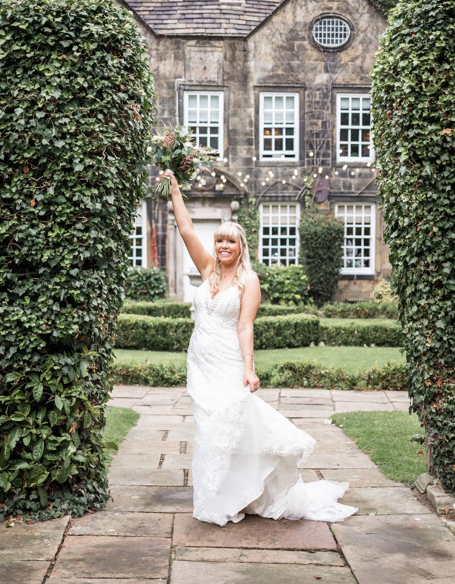 Bride in white dress poses joyfully on stone path between ivy-covered walls of historic building.