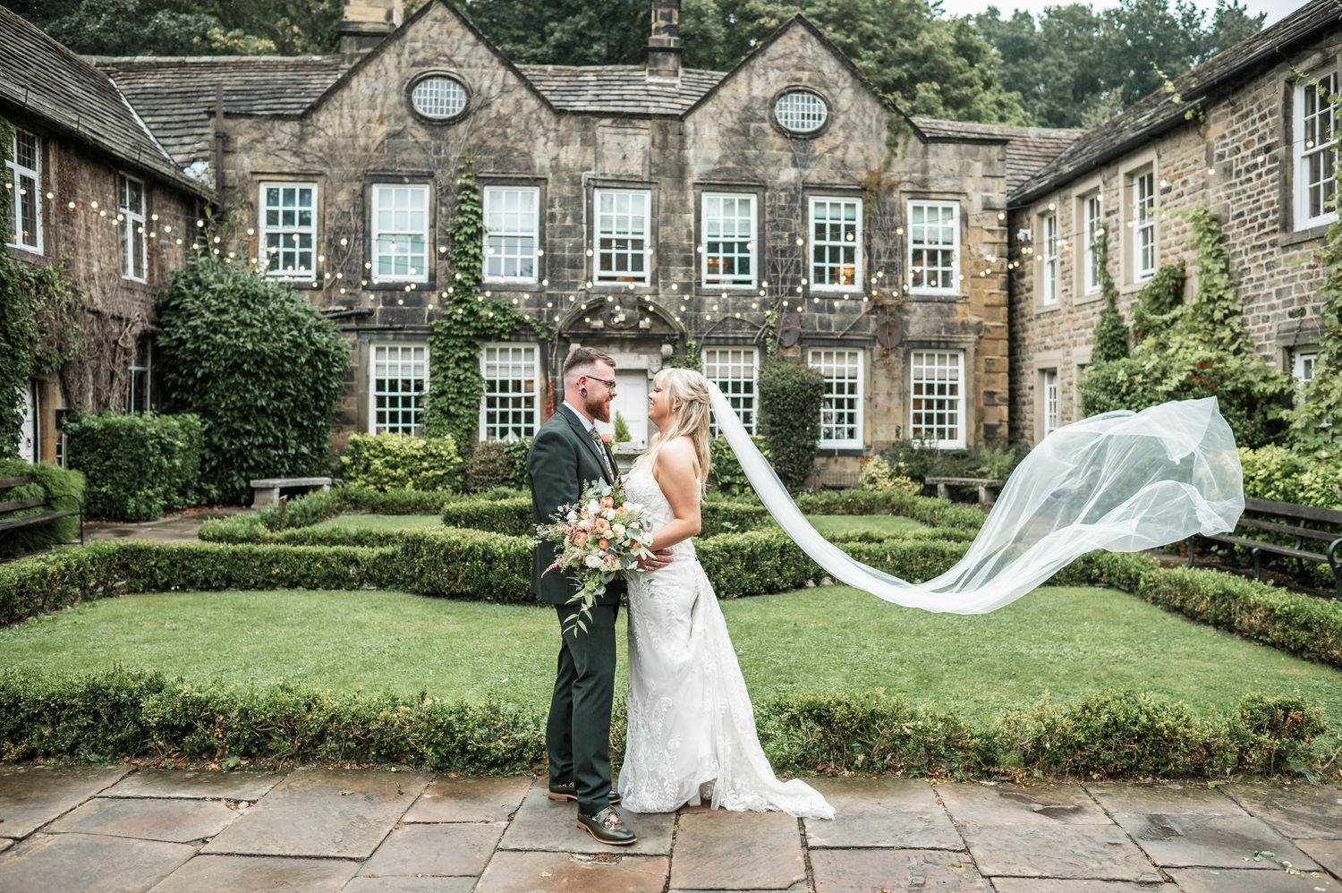 A dramatic wedding photo with flowing veil against historic stone building and manicured gardens.