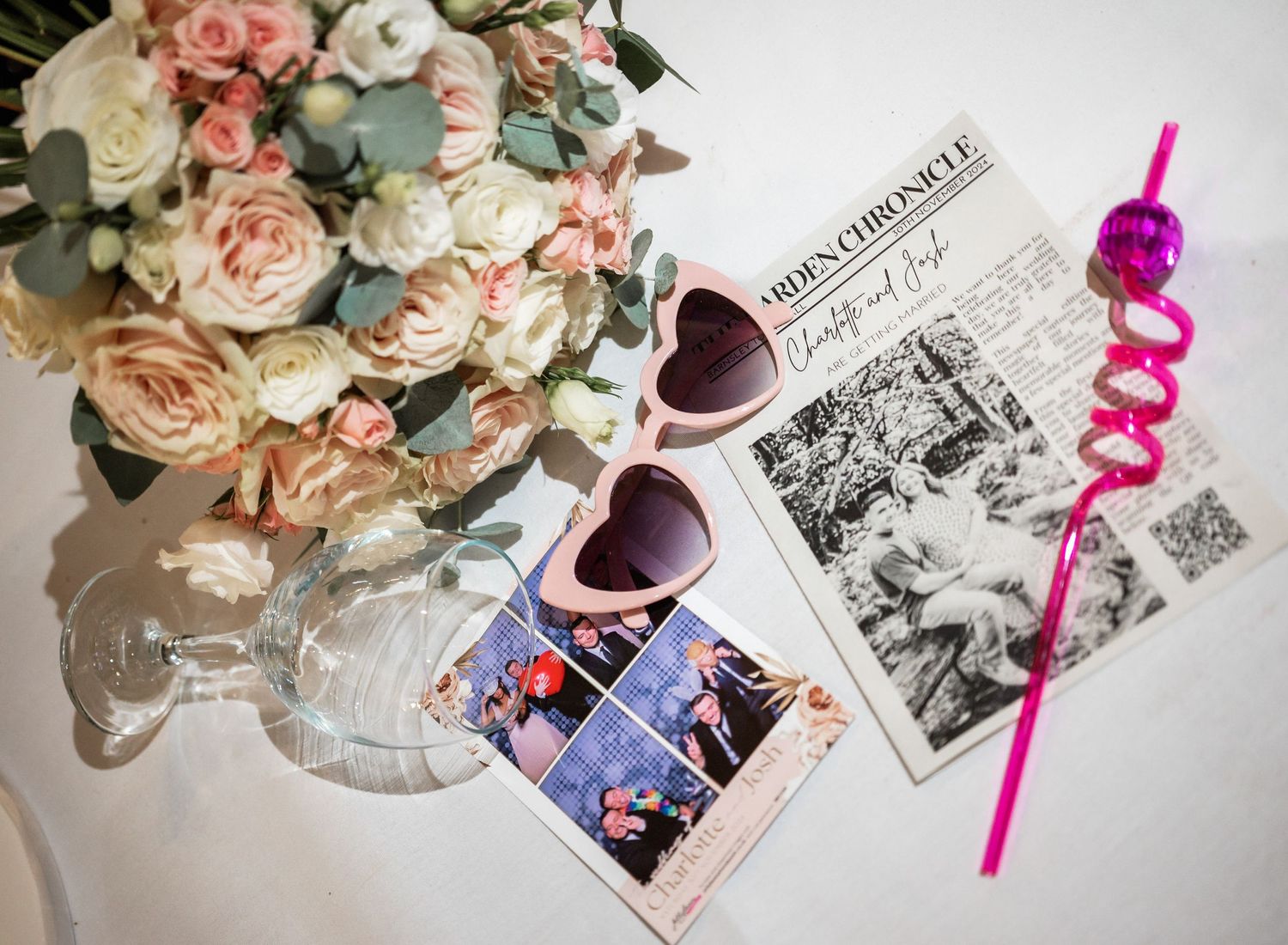 Wedding details flatlay with roses, heart sunglasses, photos and a pink straw.