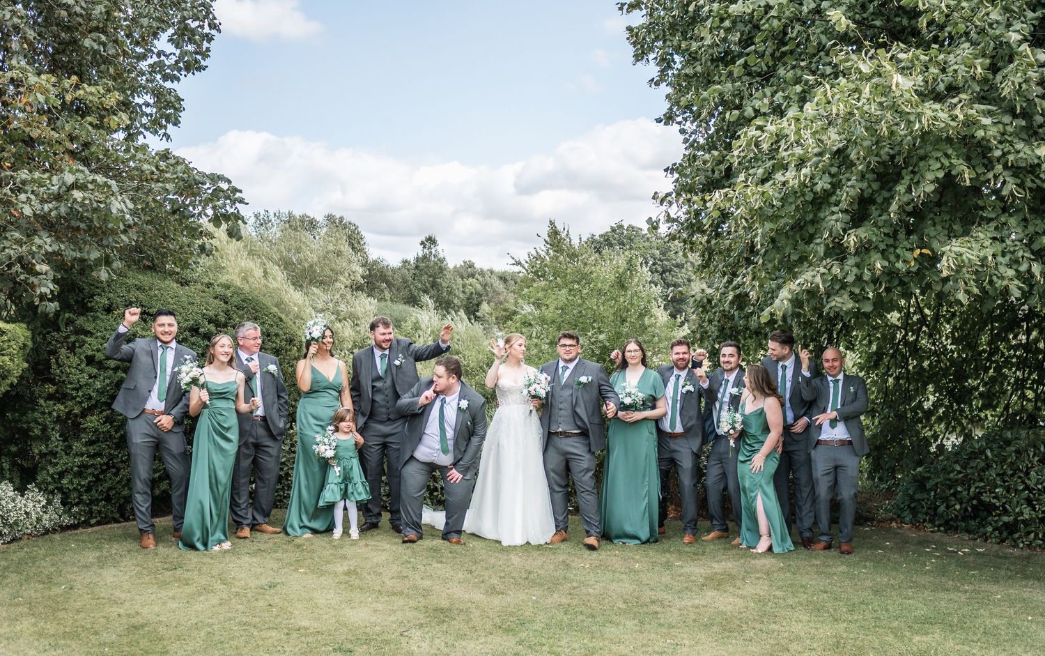 Wedding party in green dresses and gray suits pose together outdoors for group photo.