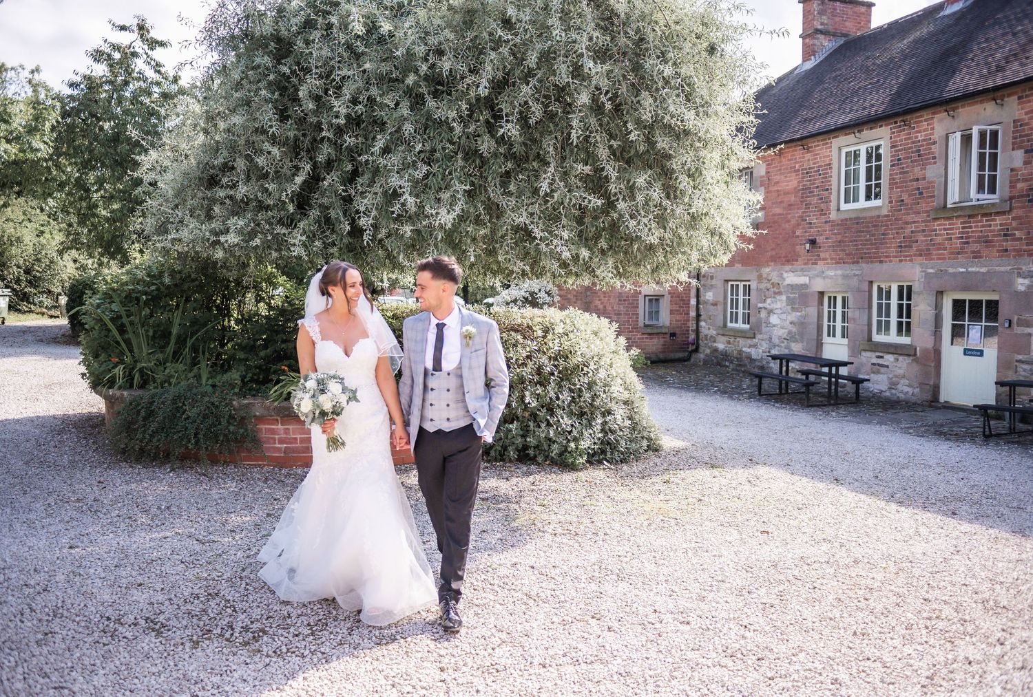 Newlyweds walk together on a gravel path outside a traditional brick country house.