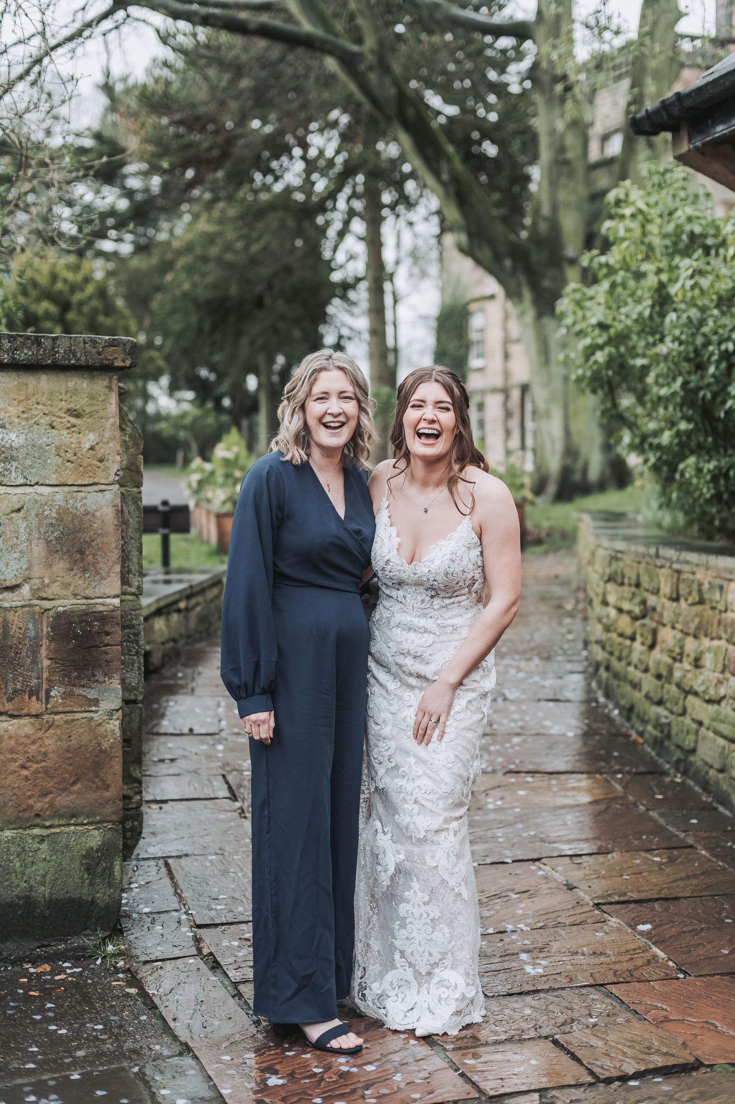 Two people share a laugh on a rainy stone pathway during wedding celebrations.