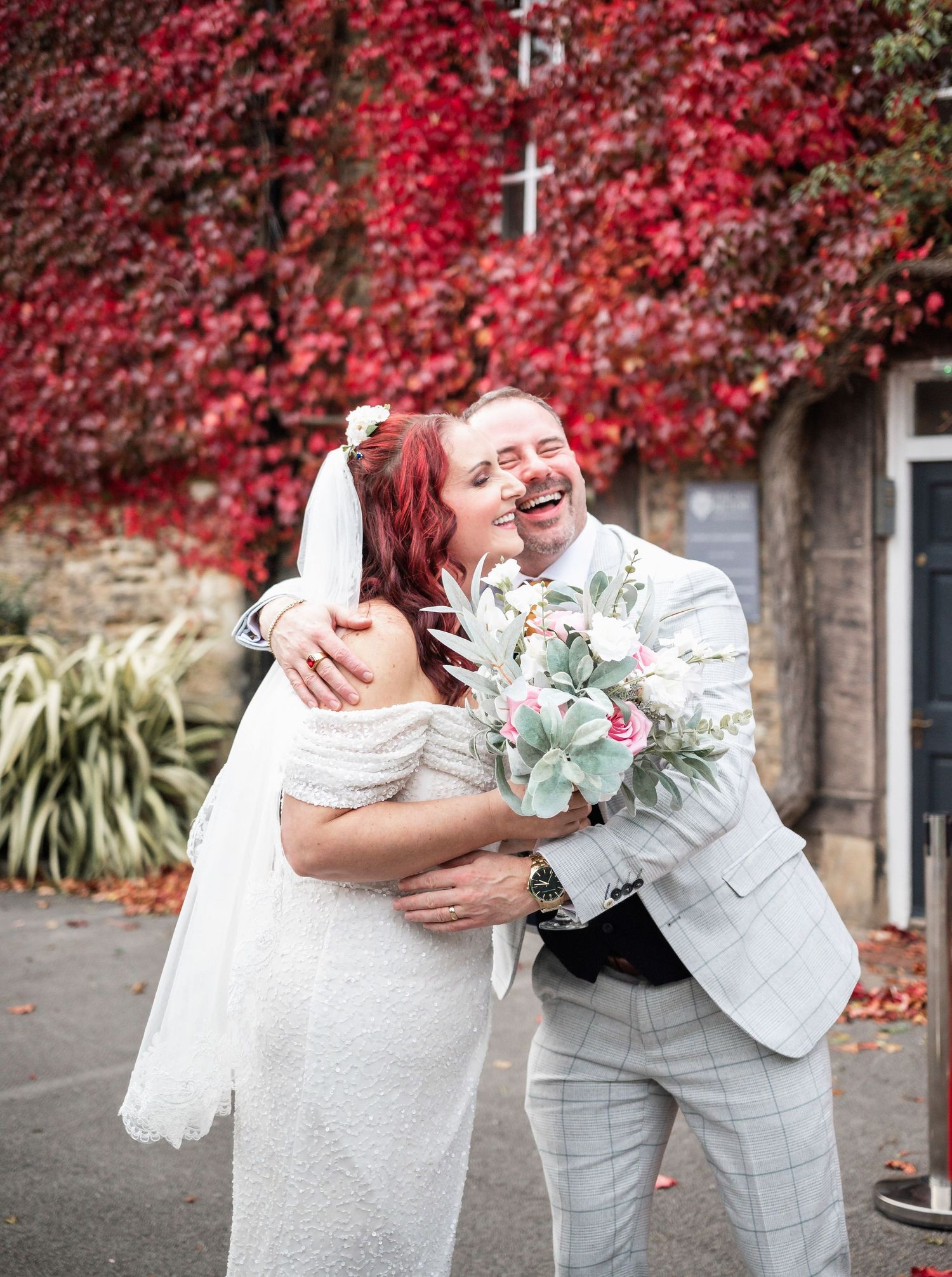 A happy wedding couple embracing outdoors against a wall of vibrant red autumn foliage.