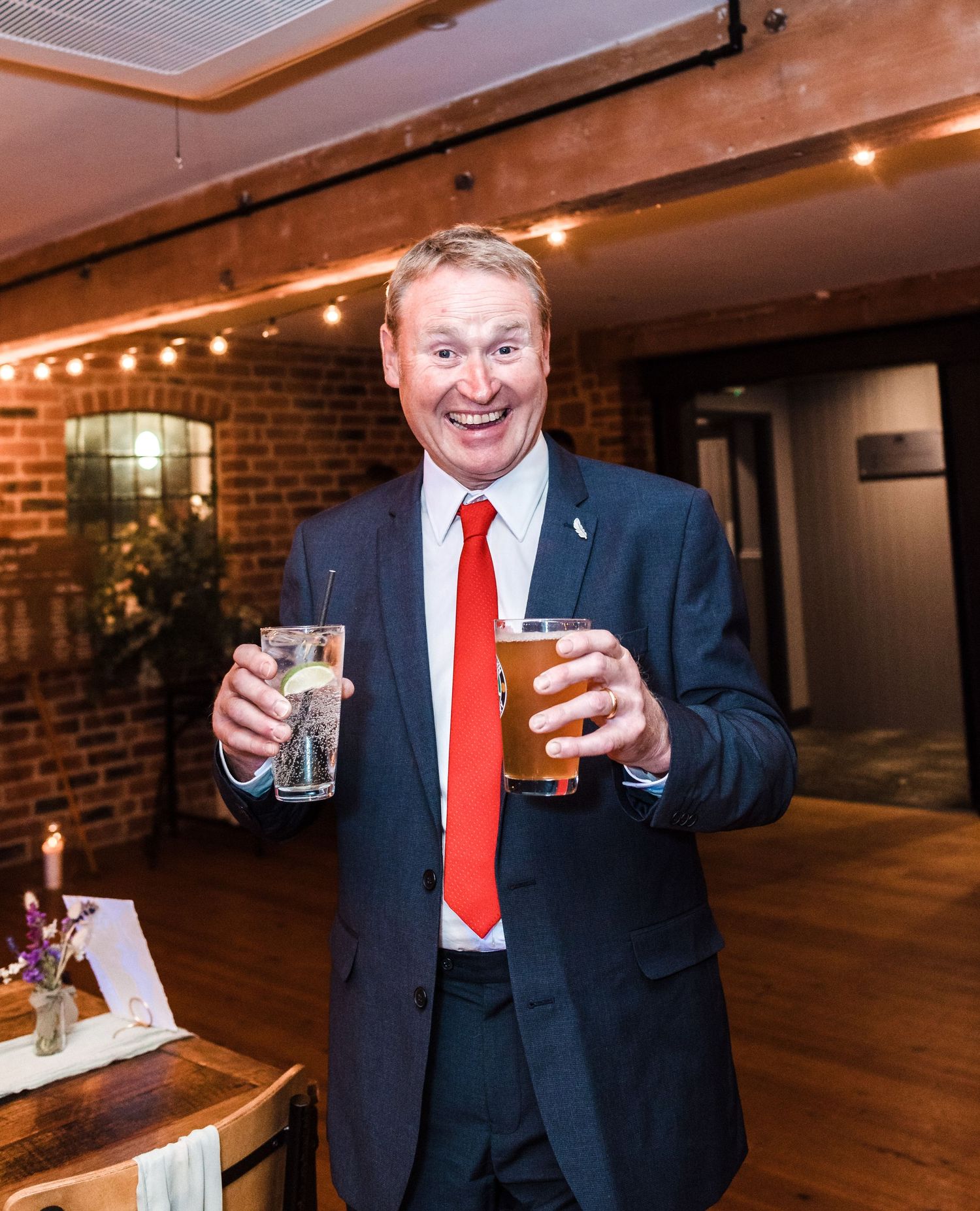 Guest in navy suit and red tie holding drinks at wedding reception.