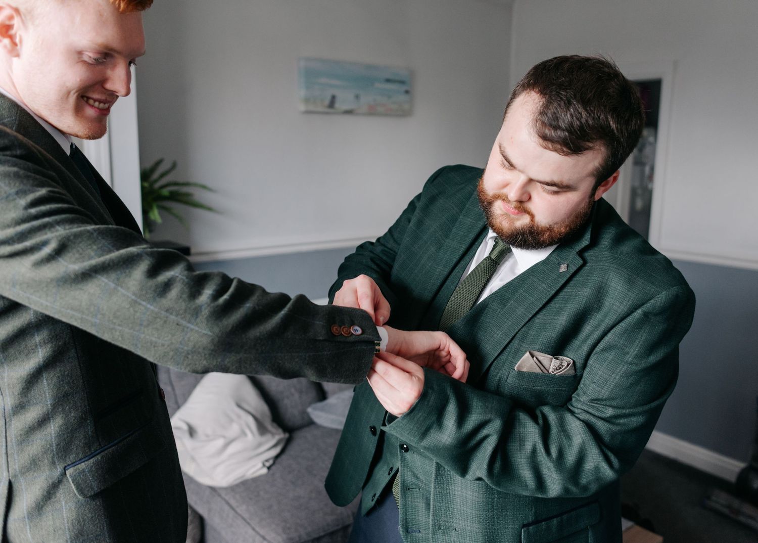 Two people in green suits adjust formal attire while getting dressed in a room with light walls.