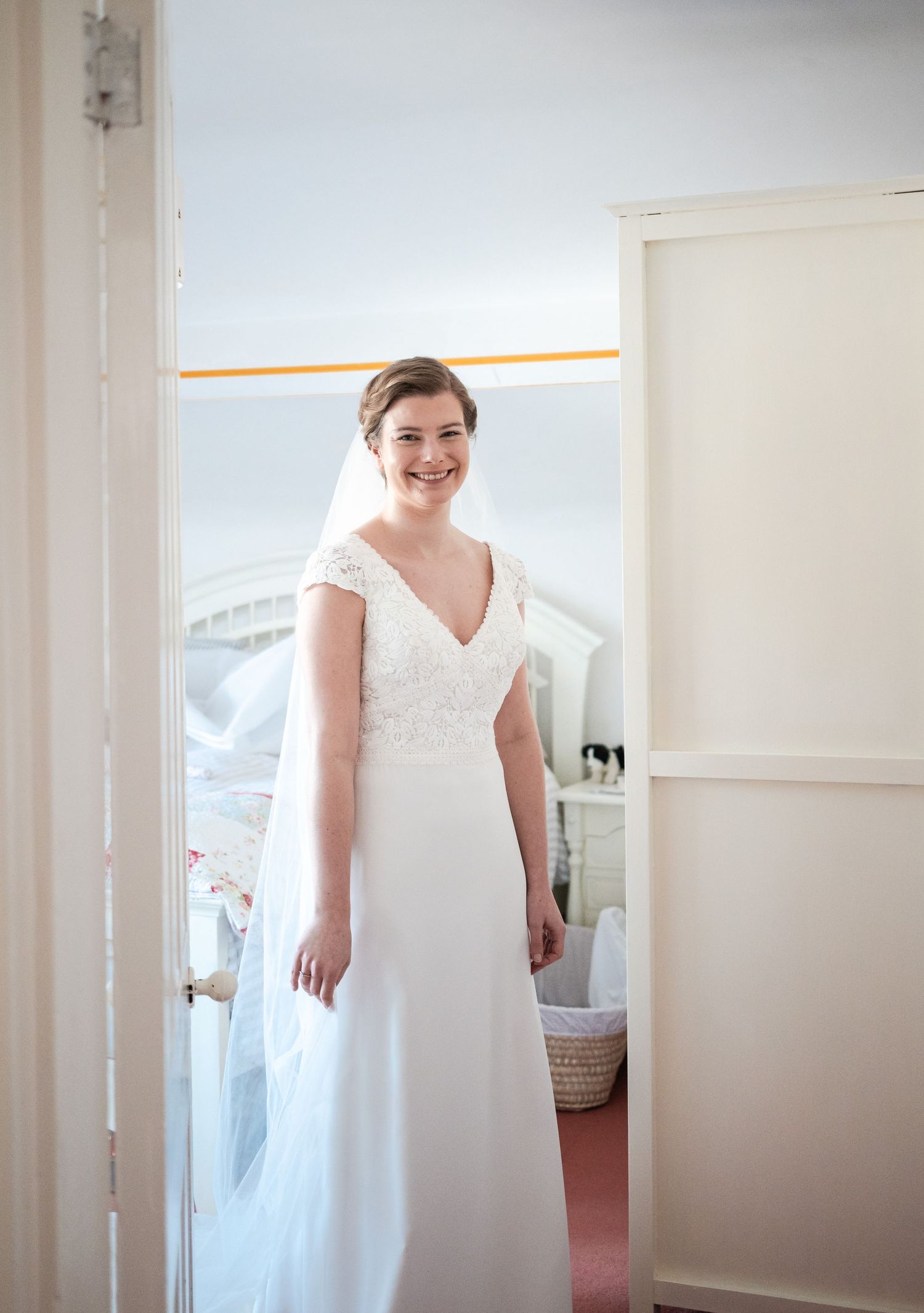 A bride in a classic white wedding dress stands in a bright room during wedding preparations.