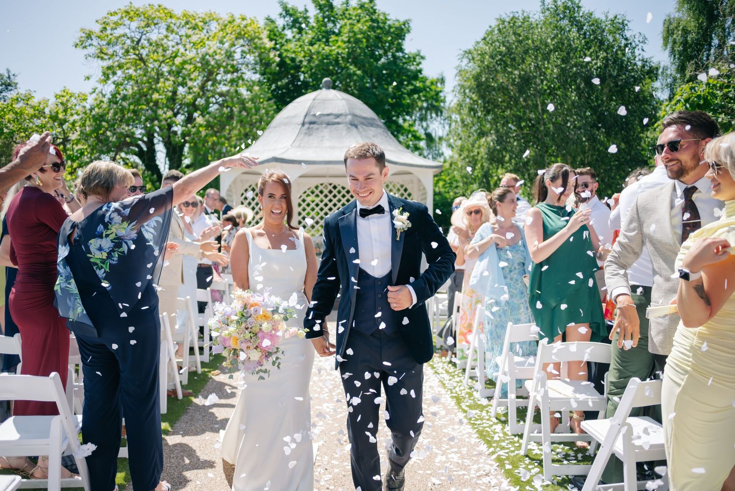 Wedding guests celebrate by throwing confetti at an outdoor garden ceremony on a sunny summer day.