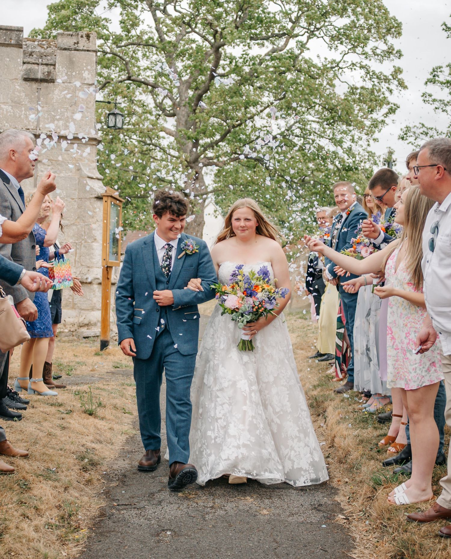 Wedding guests throw confetti over newlyweds outside historic stone church.